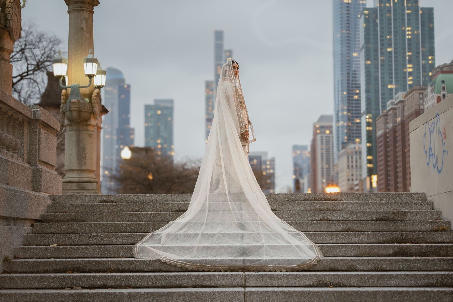 South Asian bride in ivory lehenga with cathedral veil stands on steps with Chicago skyline backdrop.