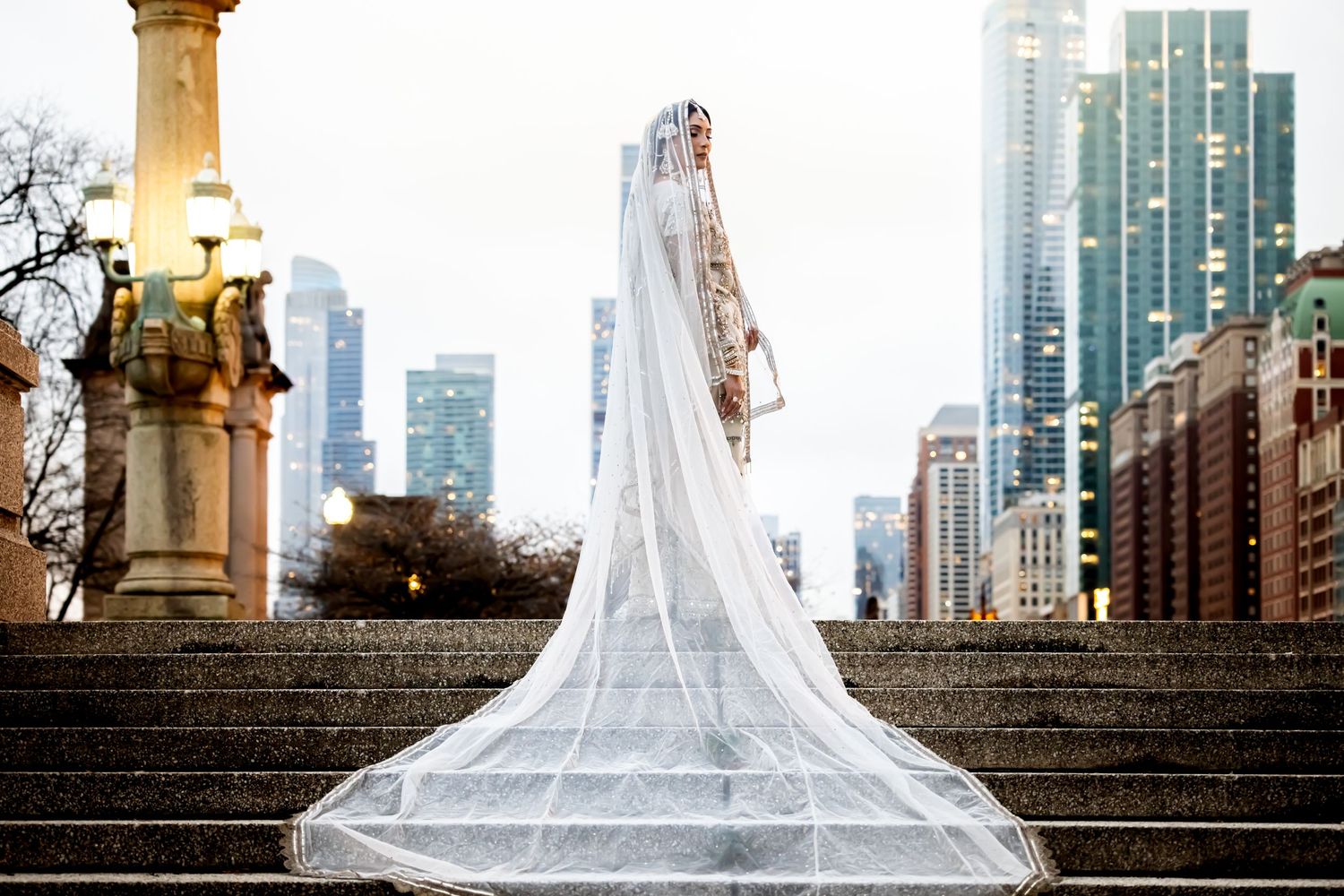 South Asian bride in embroidered ivory lehenga with dramatic cathedral veil near monument and city skyline.
