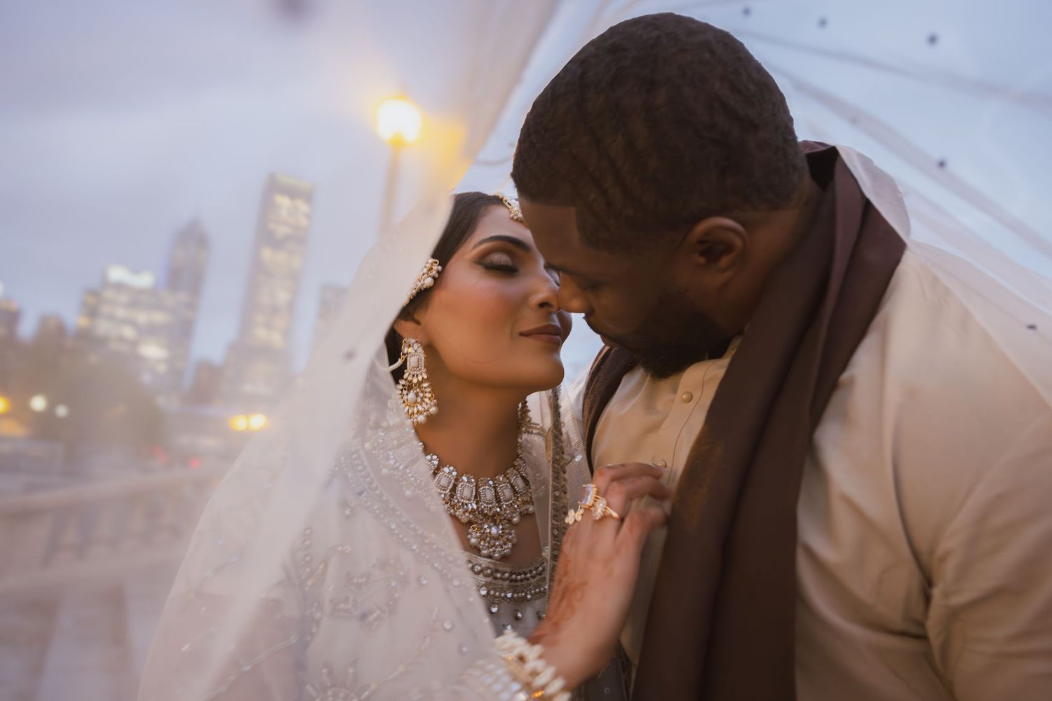 Close romantic portrait of bride in embellished hijab and groom beneath sheer wedding veil at dusk.