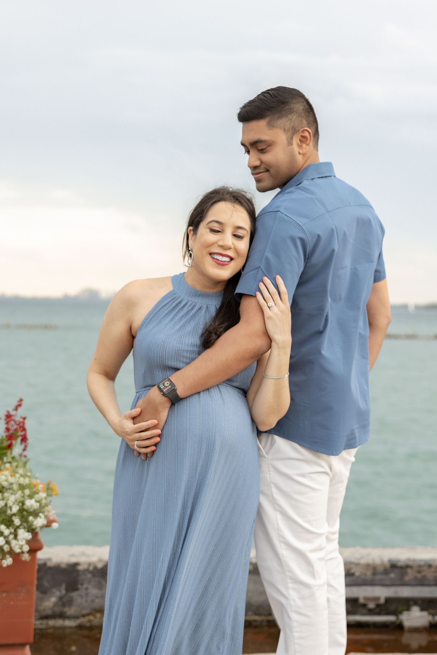 Joyful pregnant woman in blue halter dress cuddling with partner by the sea.
