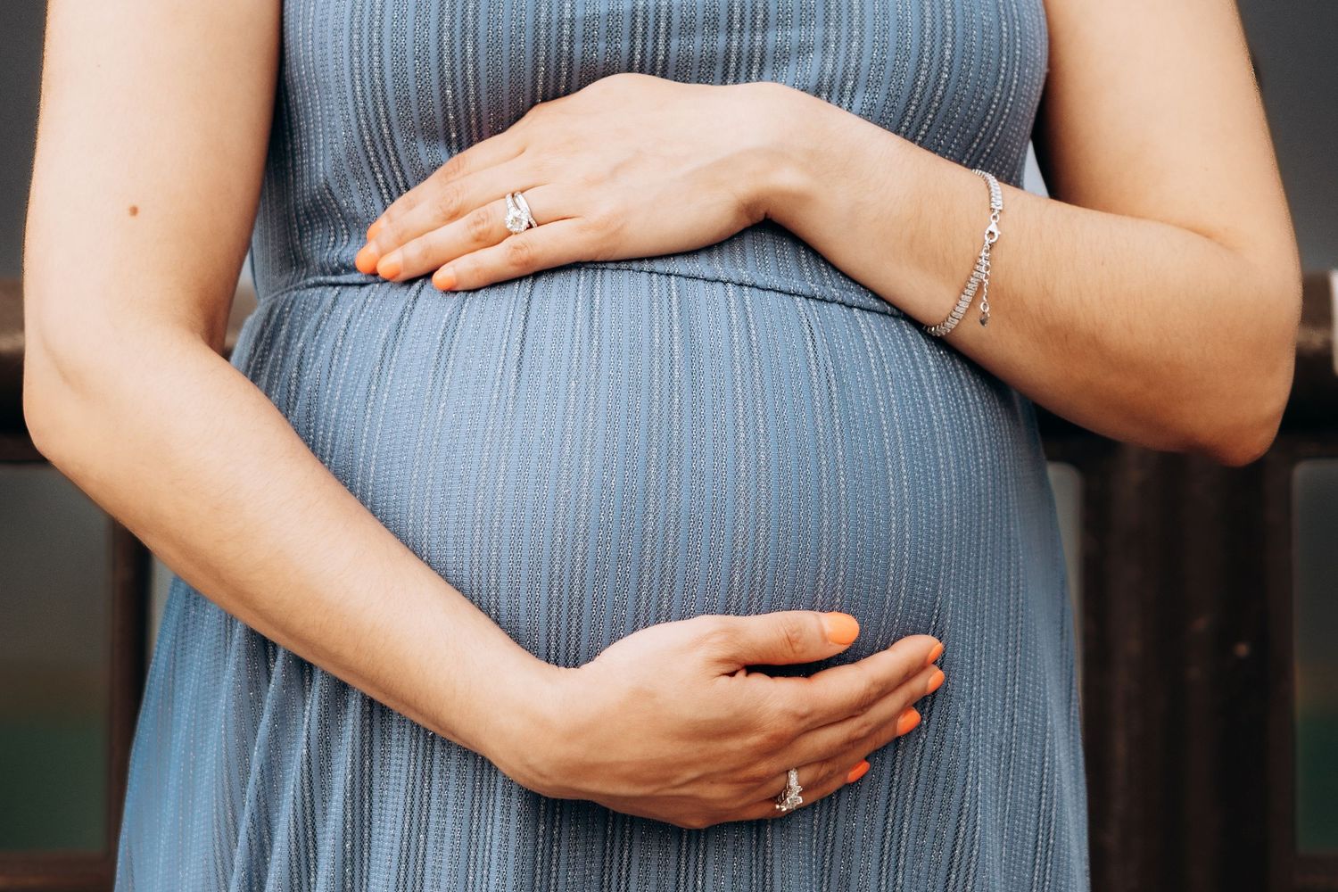 Close-up of pregnant woman in blue dress cradling her baby bump with both hands, wearing a delicate bracelet and ring.