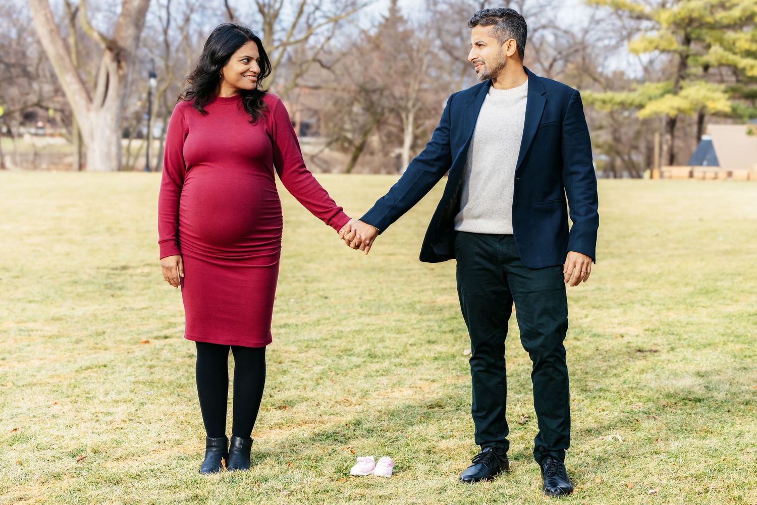 Expecting couple holding hands in a park, woman in red maternity dress smiling joyfully.