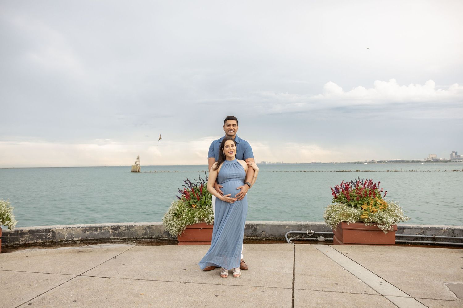 Pregnant couple standing together on waterfront promenade, woman in flowing blue dress, cloudy sky and lake backdrop.