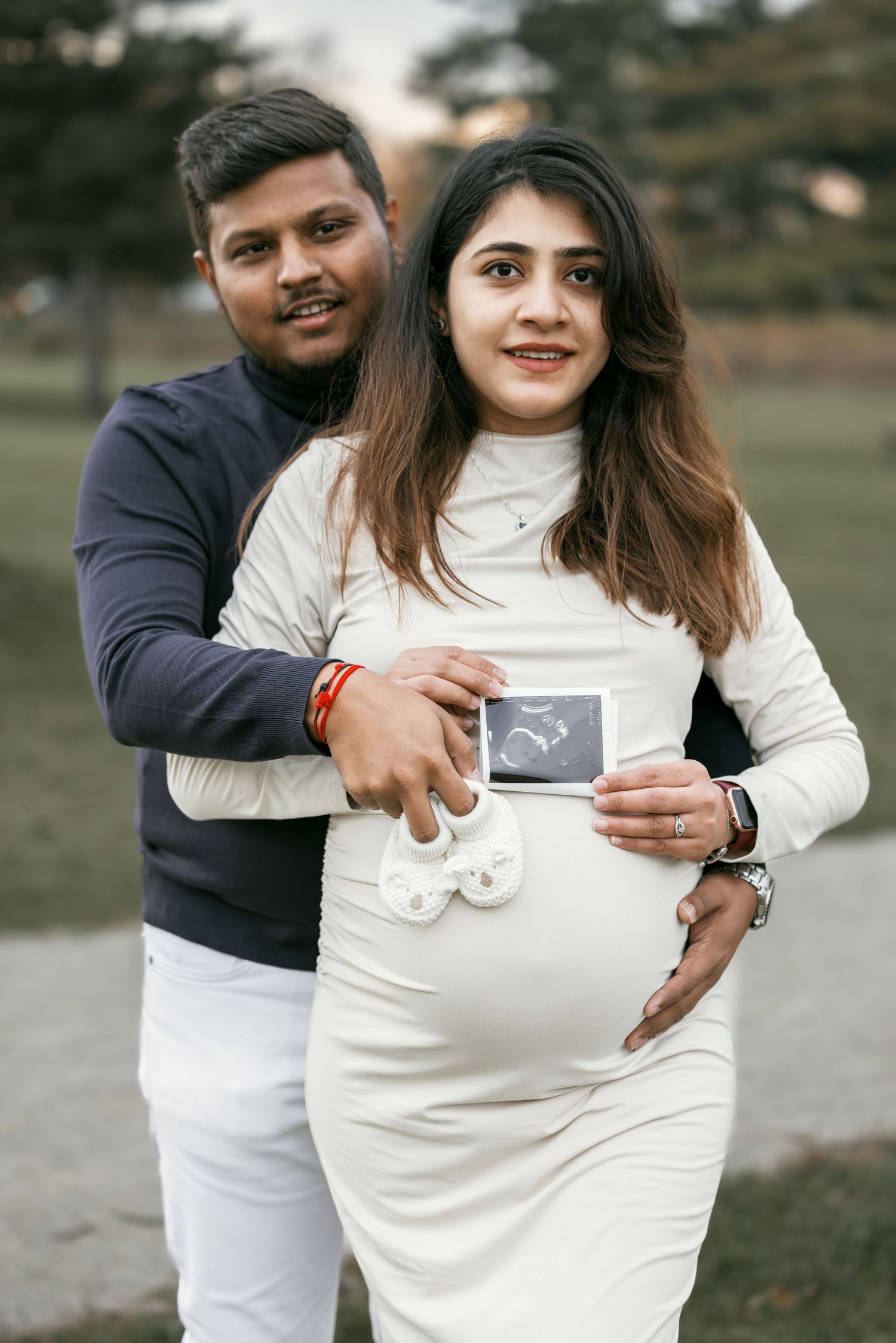 Couple holding ultrasound photo and baby shoes during outdoor maternity photoshoot.