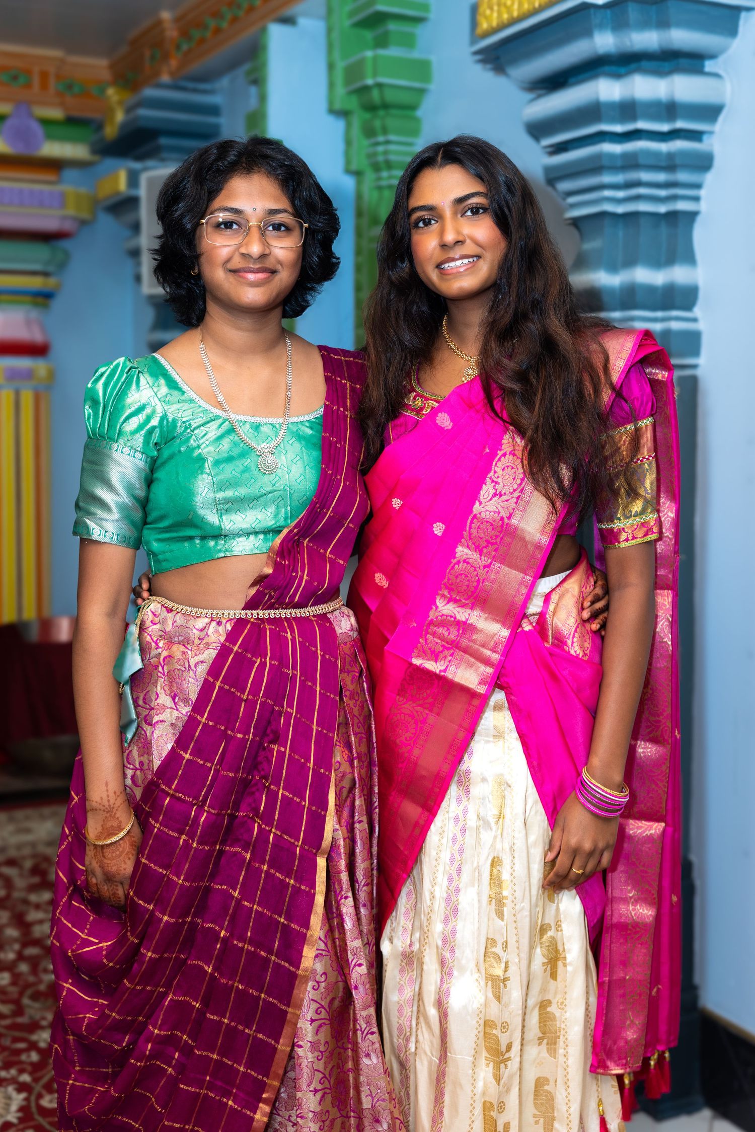 Two young Indian women in colorful traditional lehenga and saree smile together in temple.