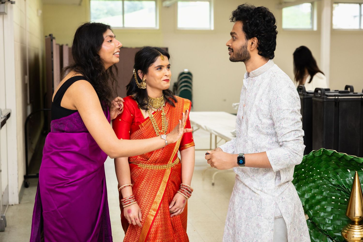 Indian bride in red saree with gold jewelry greets guests at traditional wedding celebration indoors.