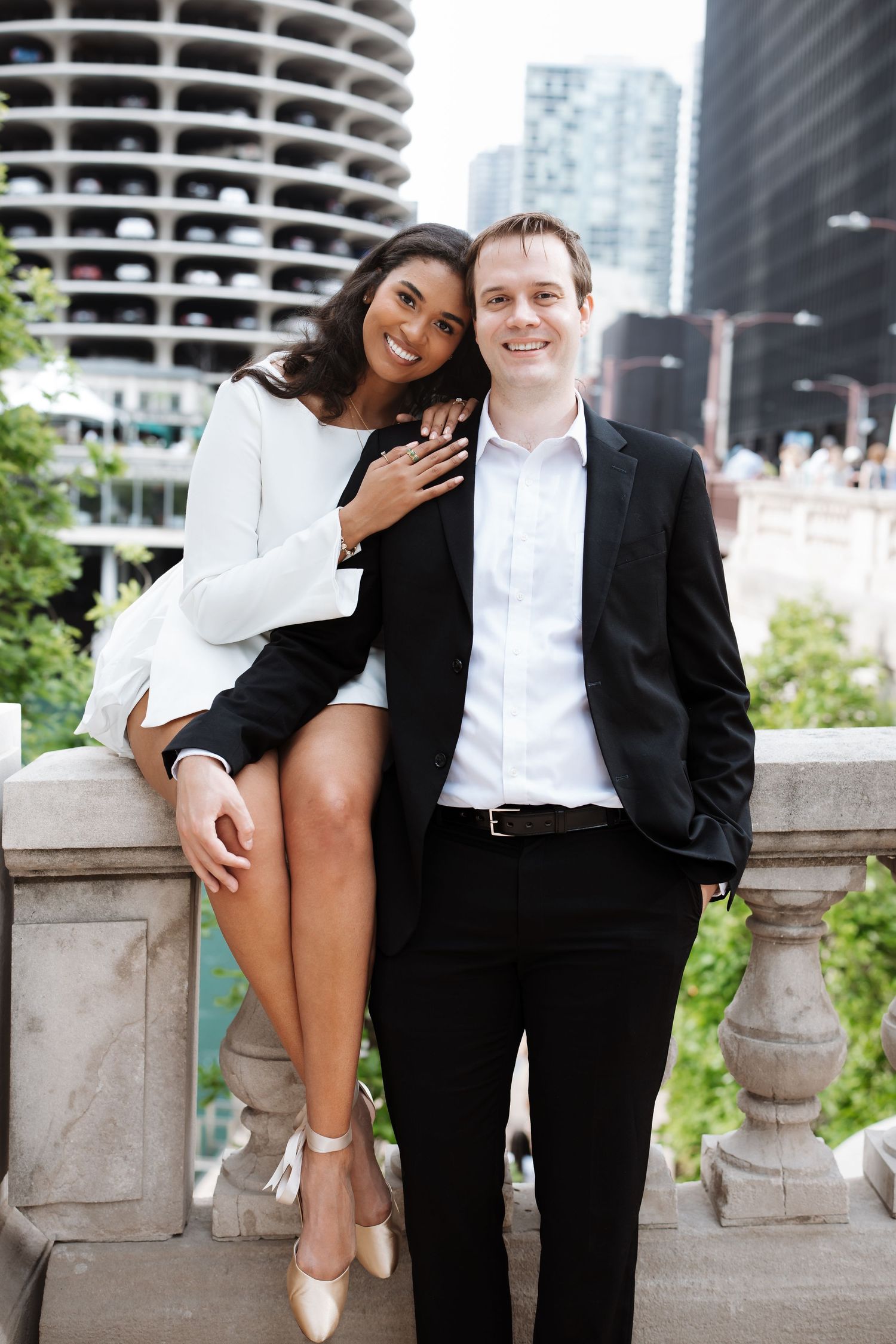Smiling interracial engaged couple posing on a stone bridge with Chicago Marina City towers in background.