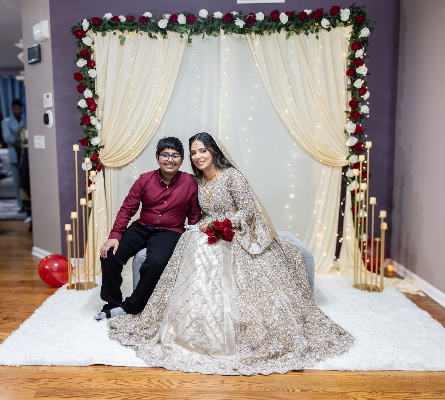 Bride in gold embroidered lehenga holds red roses posing with young boy at floral wedding backdrop.