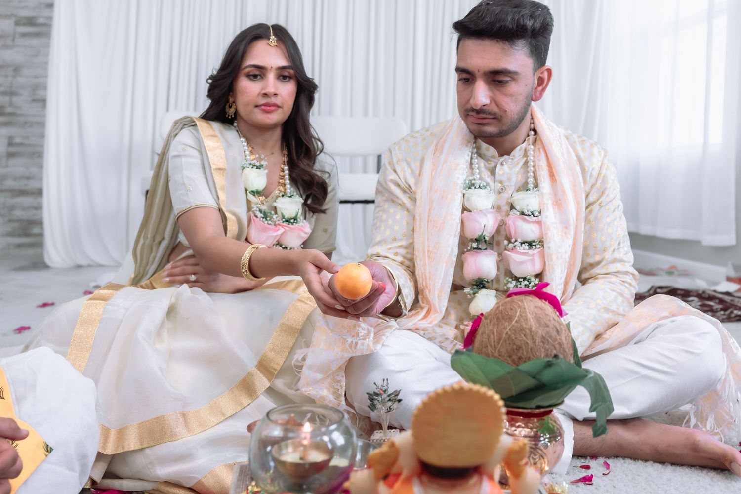 Indian couple in traditional attire perform wedding ritual seated on floor with offerings and candles during ceremony.
