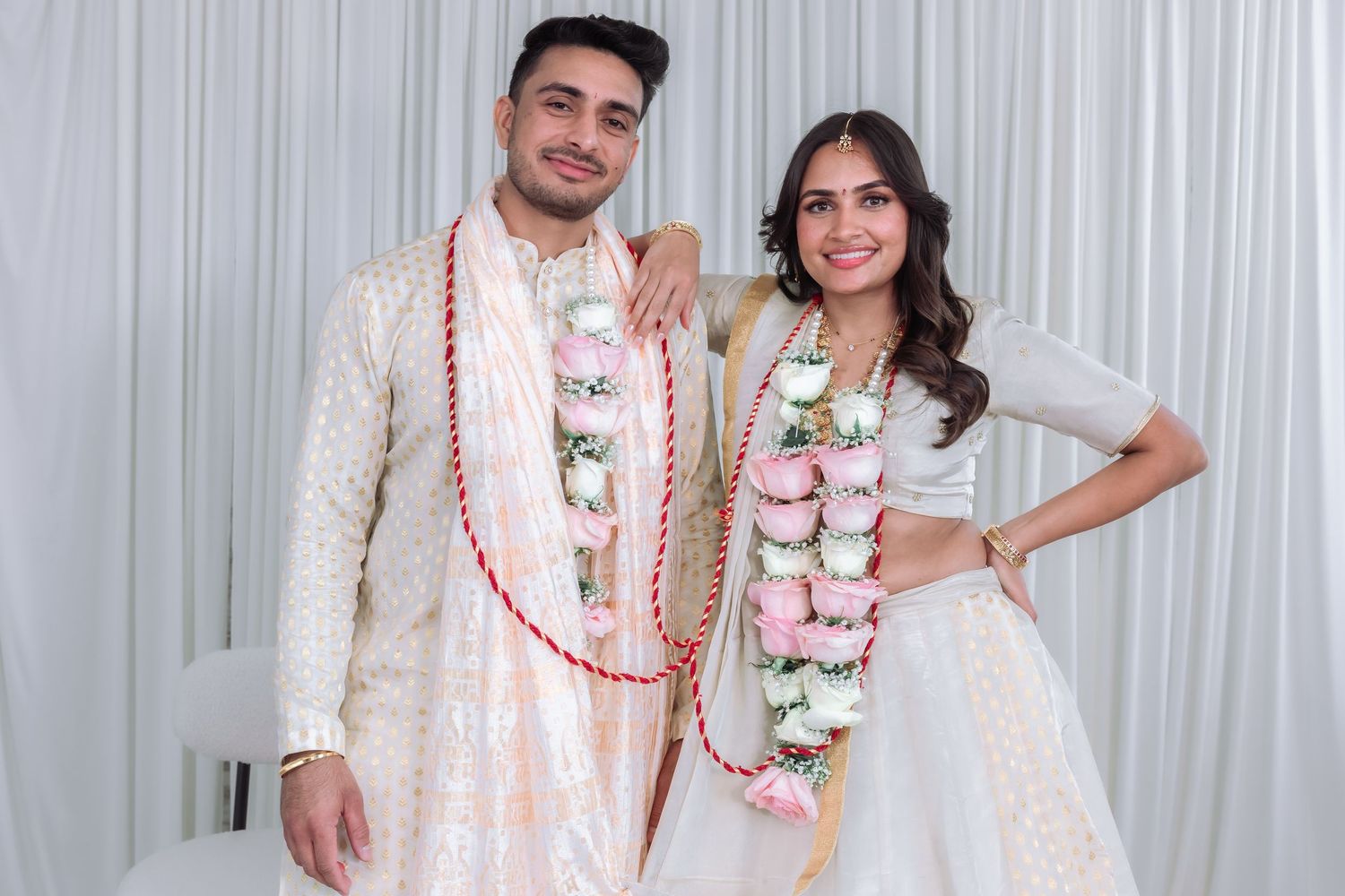 Indian bride and groom in white and gold traditional wedding outfits smile together wearing red and pink floral garlands.