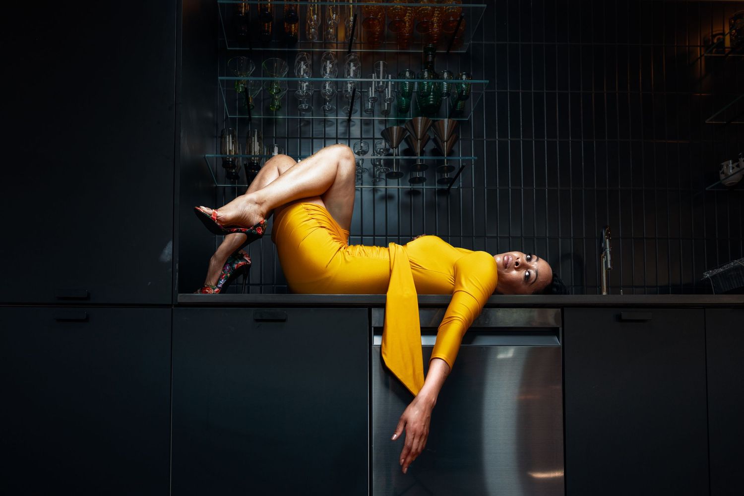 Woman in bright yellow dress poses dramatically lying across dark kitchen counter top.