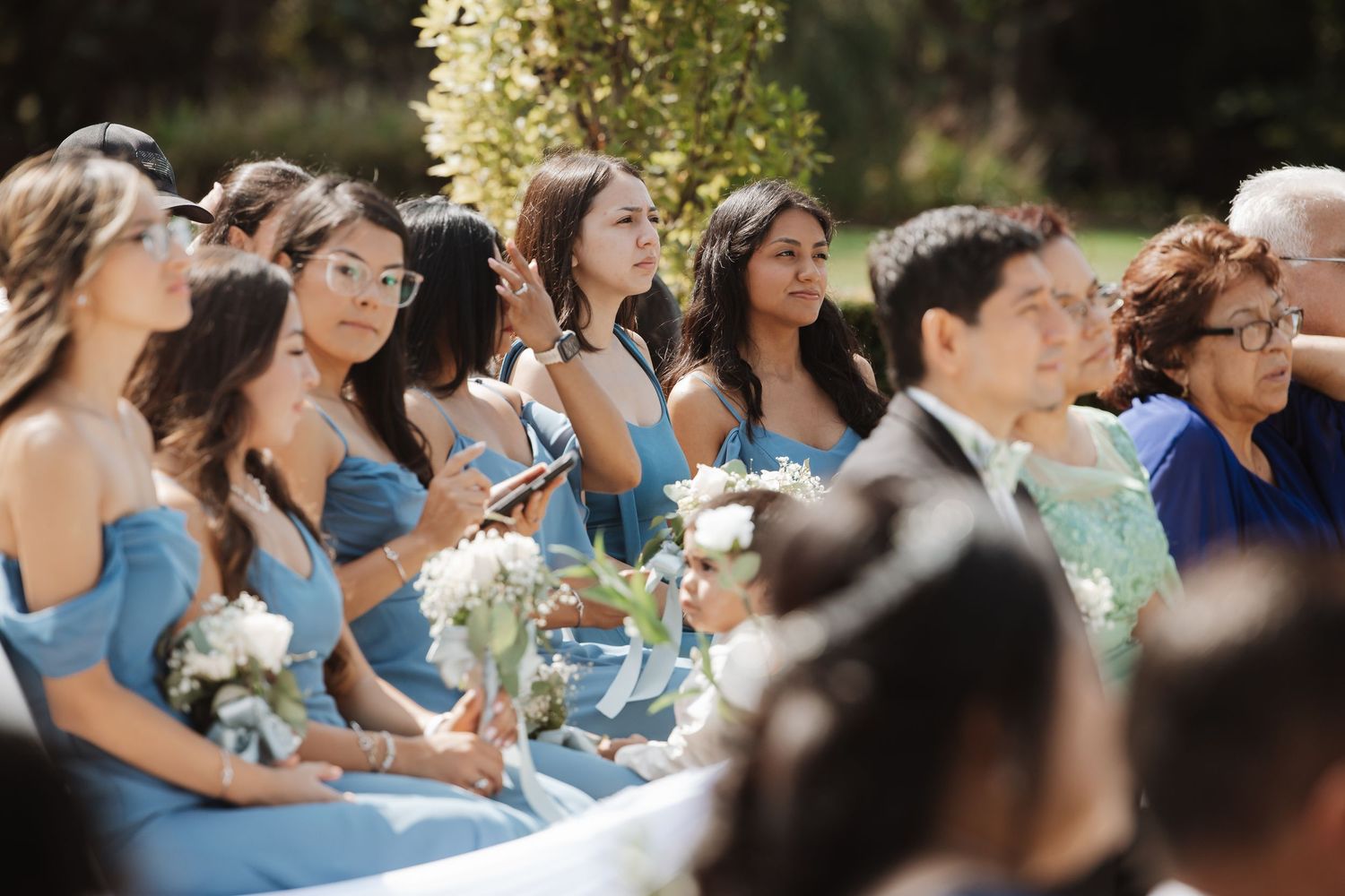 Bridesmaids in light blue dresses holding white bouquets seated at an outdoor wedding ceremony.