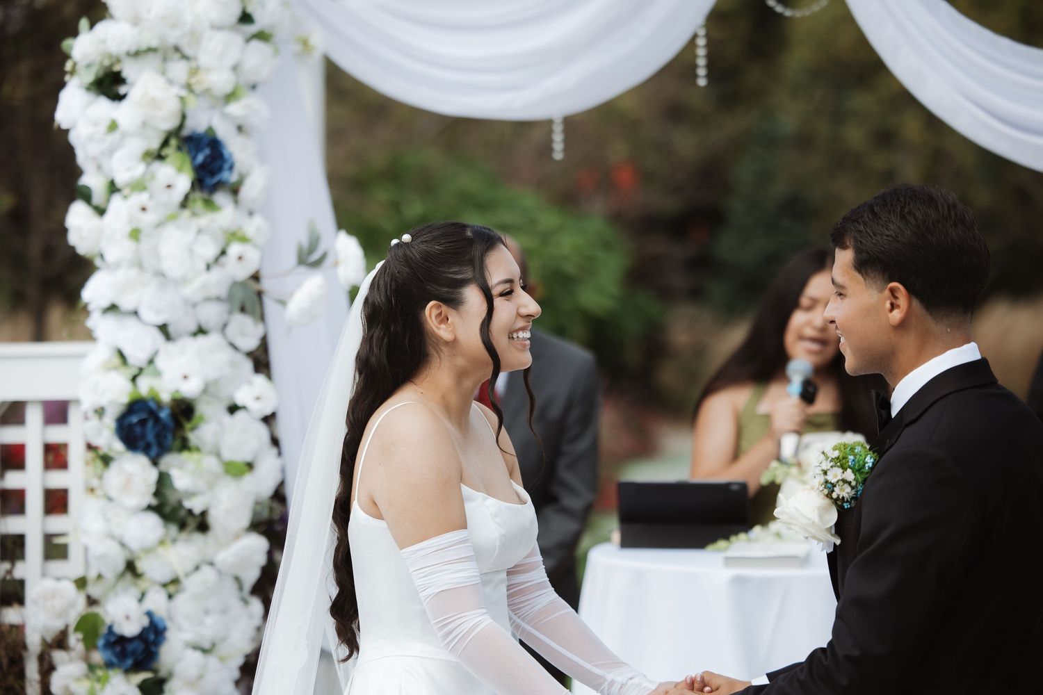 Smiling bride and groom hold hands at outdoor wedding ceremony with white floral arch.