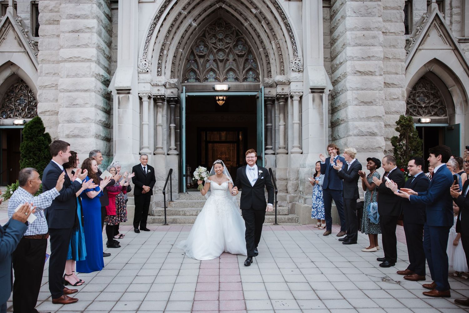 Newlyweds walk through cheering guests outside ornate gothic stone church after their wedding ceremony.