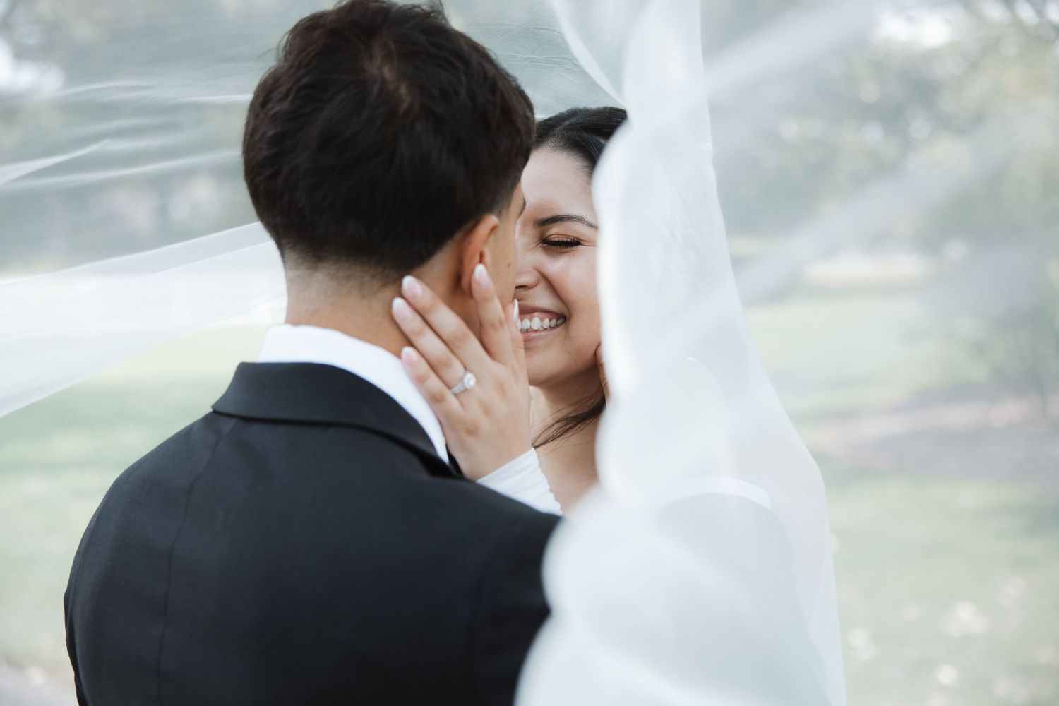 Radiant bride smiles while embracing groom under a flowing white bridal veil in an outdoor setting.
