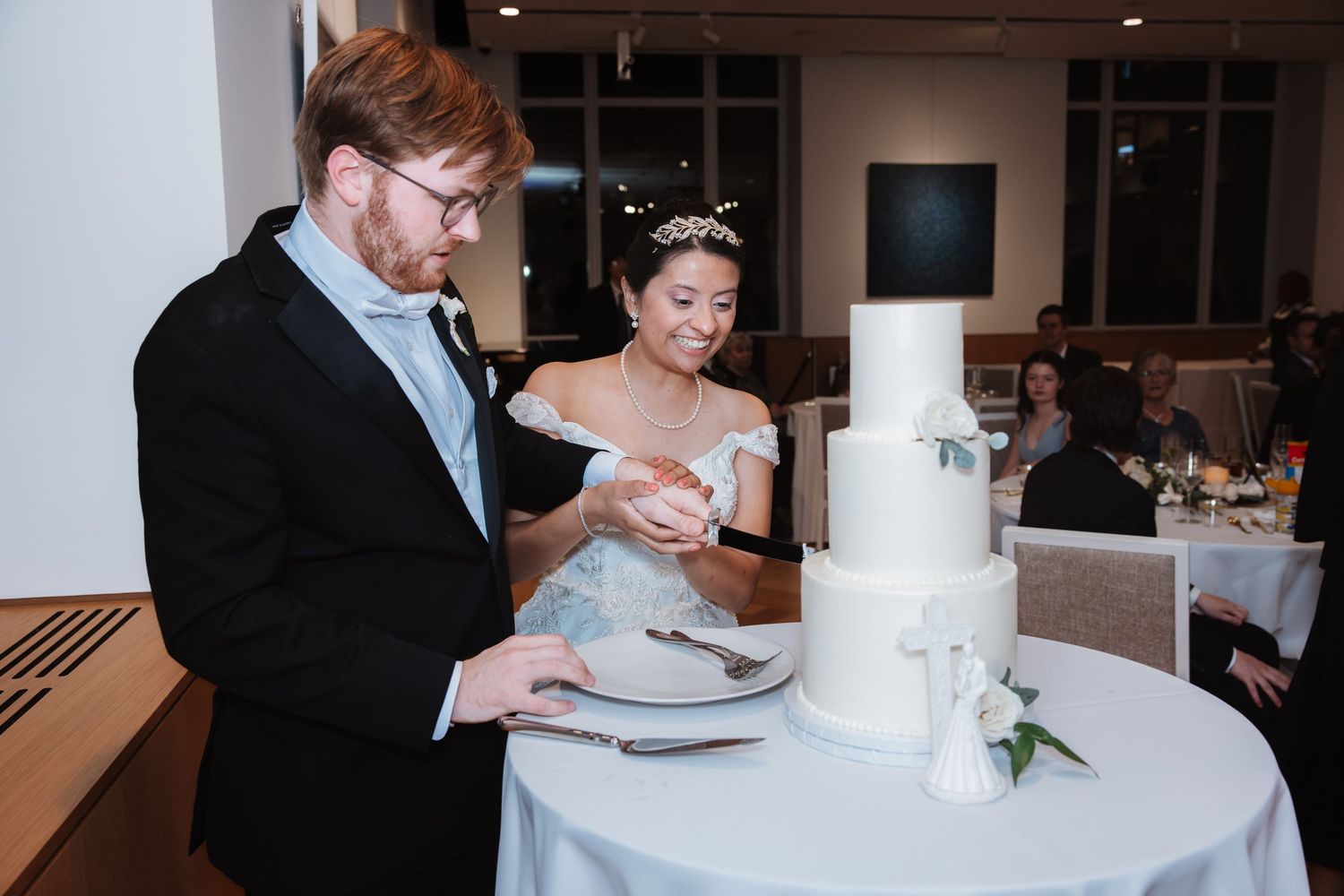 Smiling bride and groom cutting elegant white three-tier wedding cake at reception dinner table.