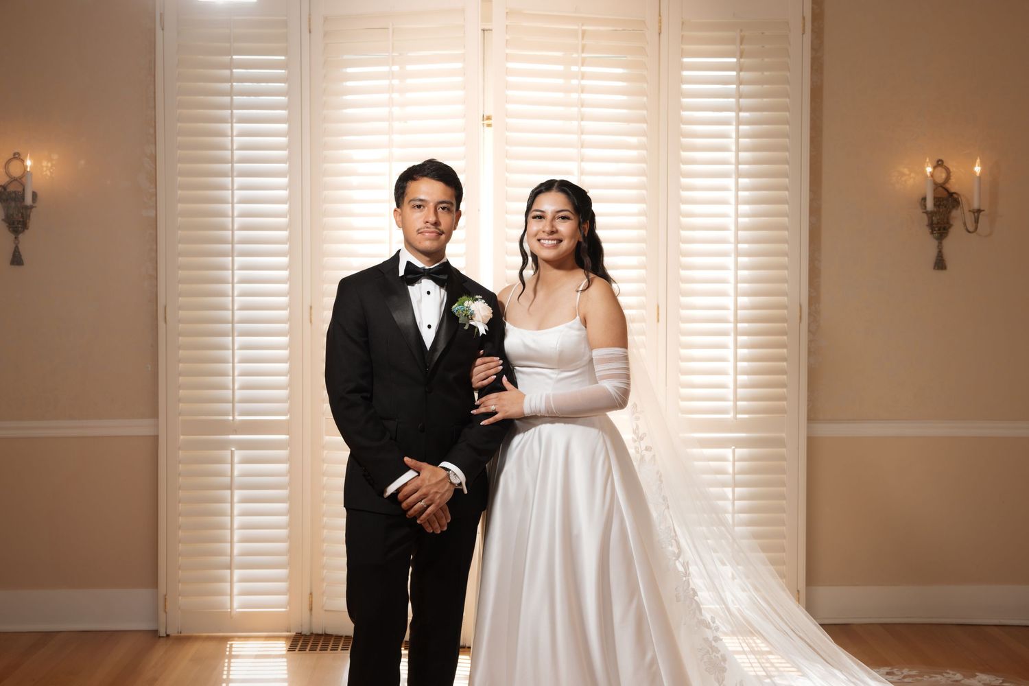 Elegant bride and groom portrait indoors, bride in white gown with long gloves, groom in classic black tuxedo.