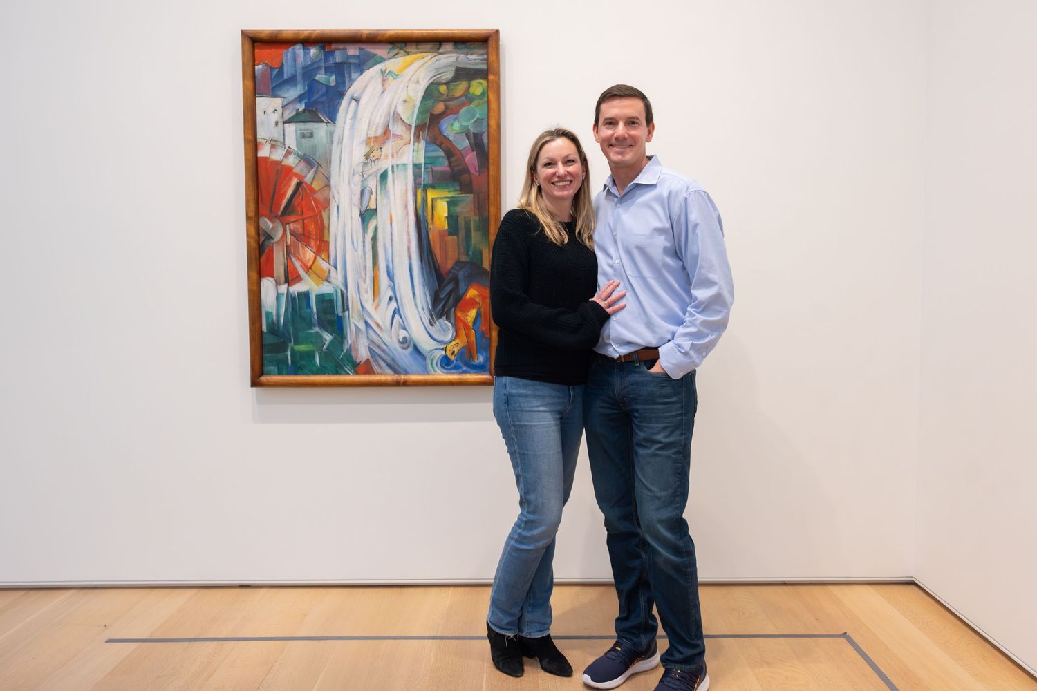 A smiling couple poses together in front of a colorful abstract painting at an art museum.