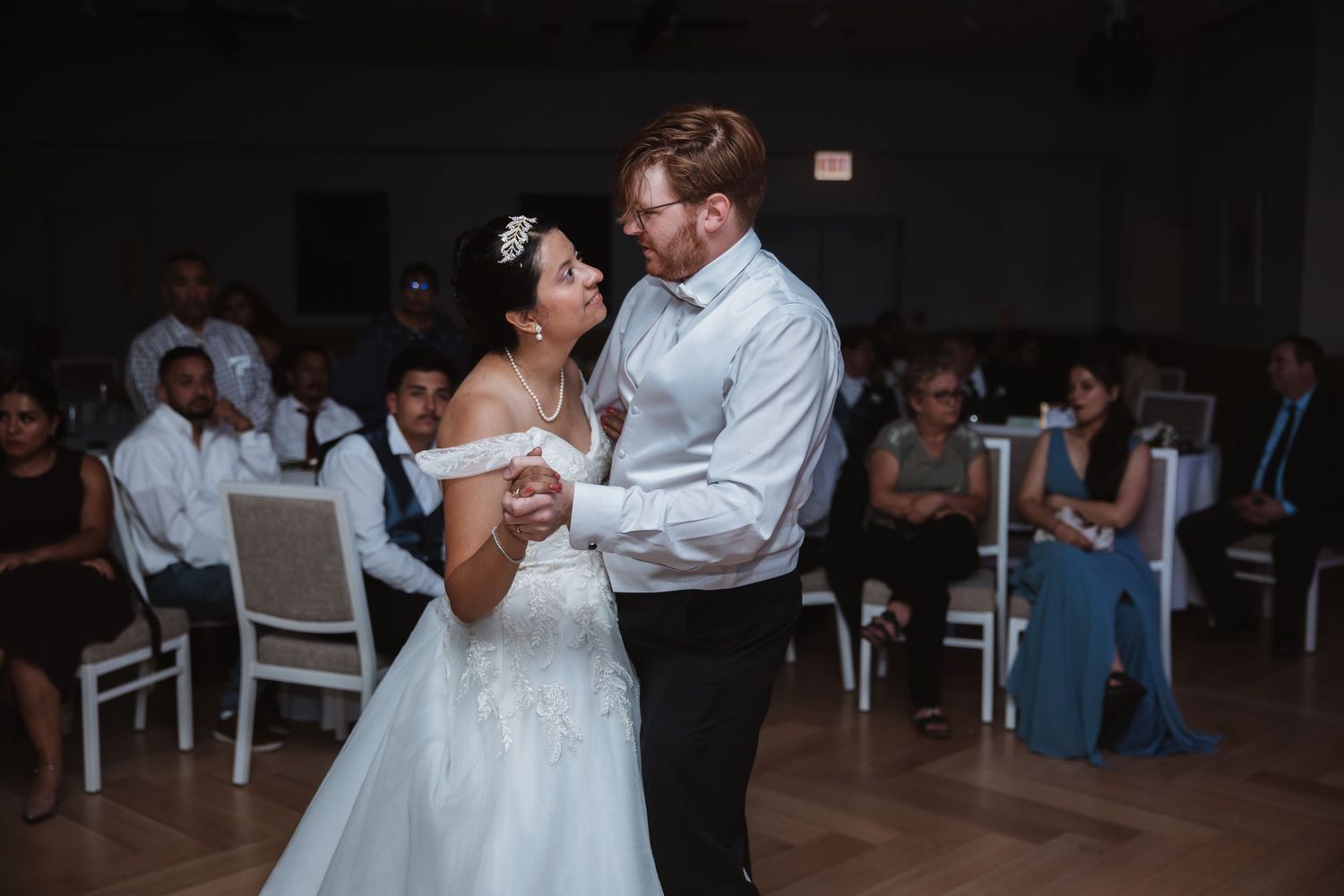 Newlyweds sharing an intimate first dance together in elegant white reception hall surrounded by wedding guests.