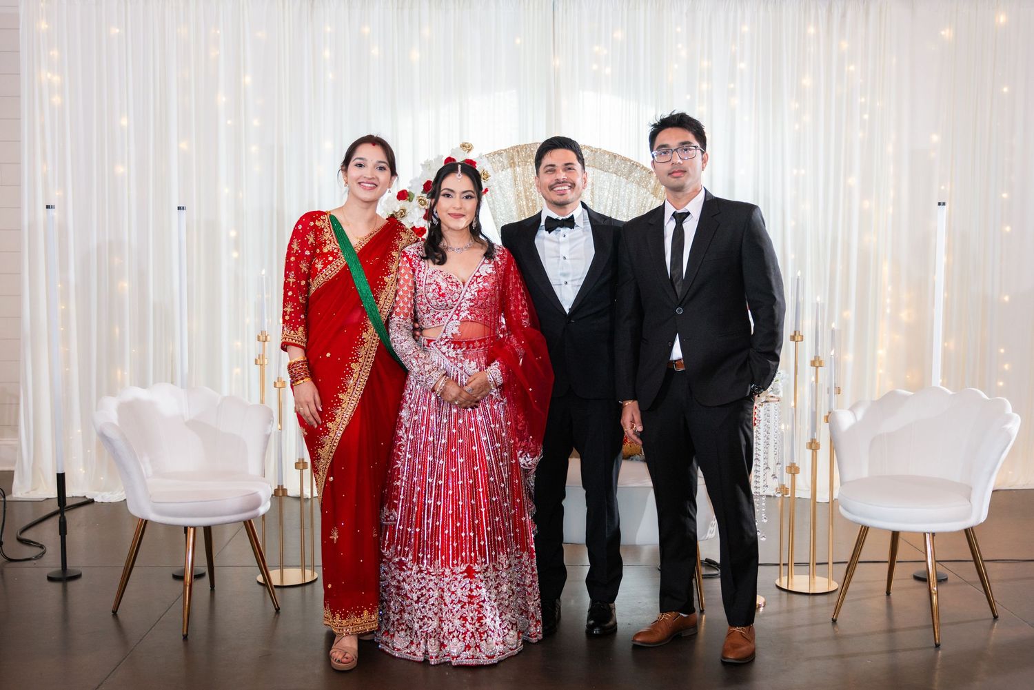 Indian wedding group photo with bride in red bridal lehenga, groom in tuxedo, and two guests in formal attire.