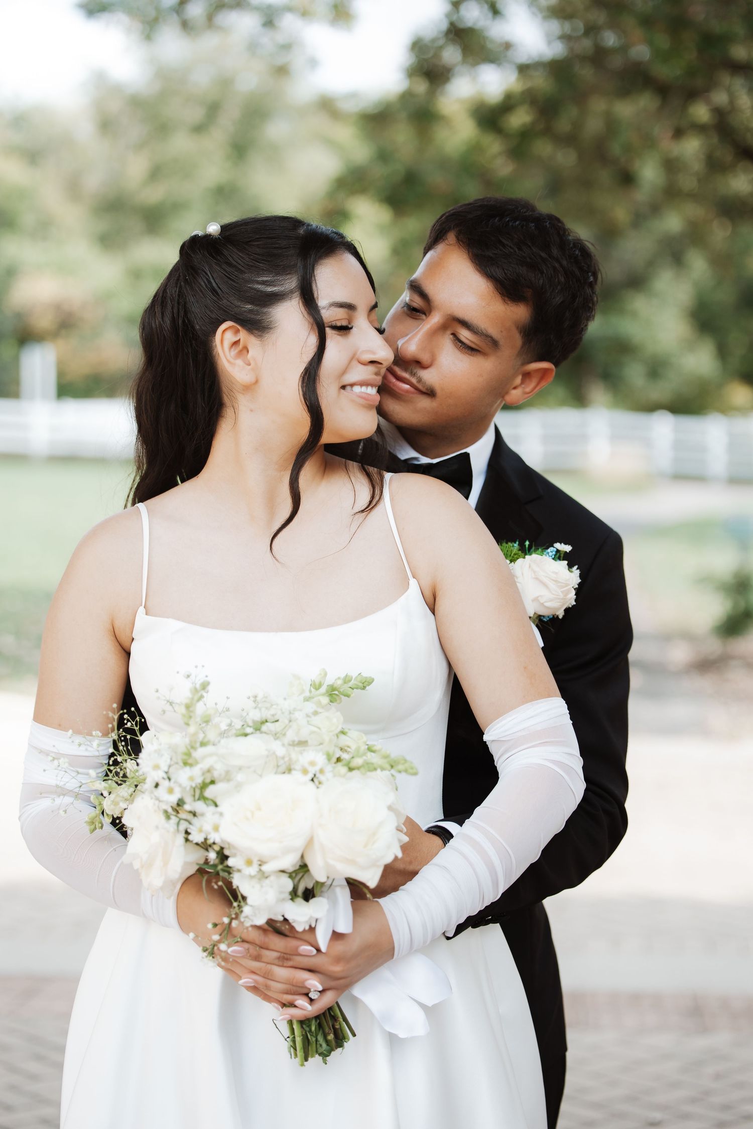 Groom in black tuxedo kissing smiling bride's cheek outdoors, bride holding white bouquet.