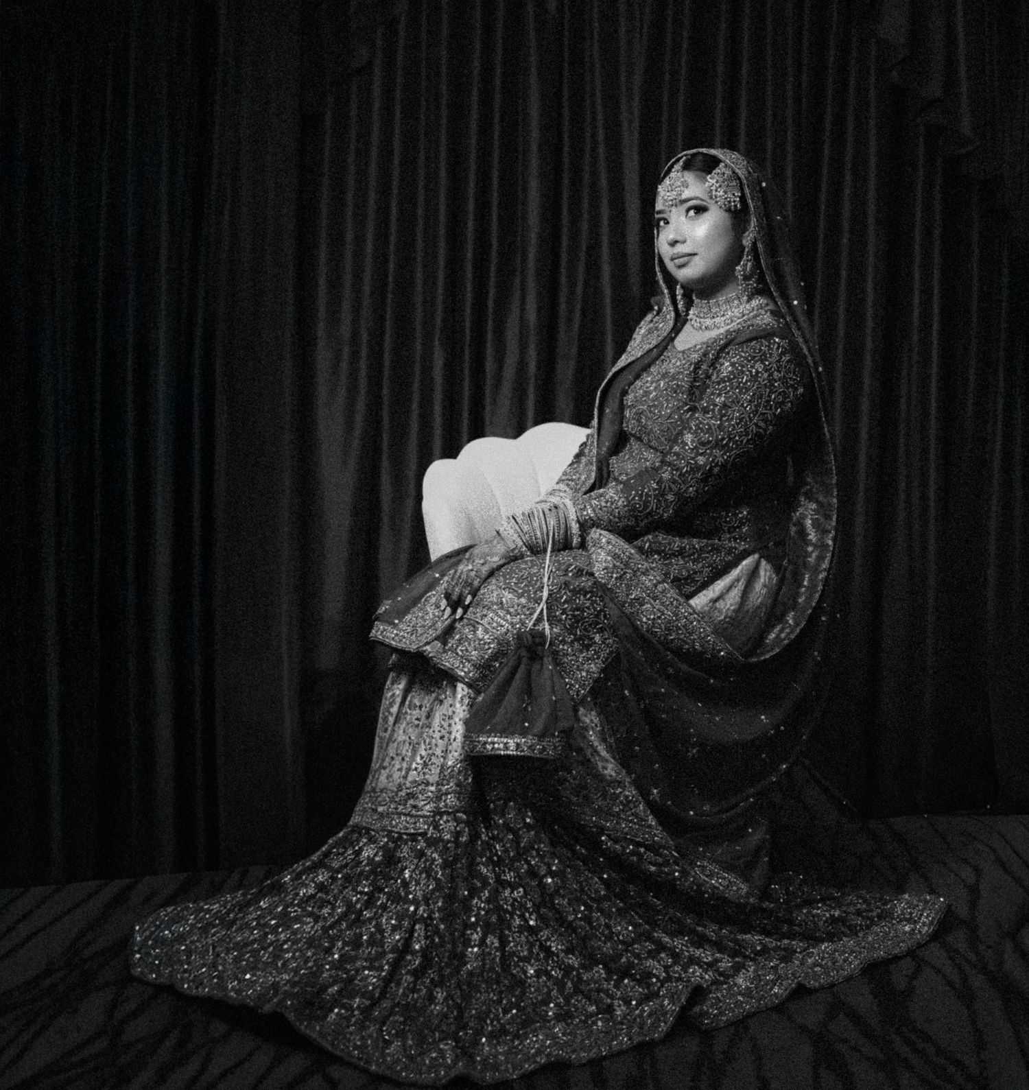Bride in ornate traditional South Asian bridal attire seated in black and white portrait.
