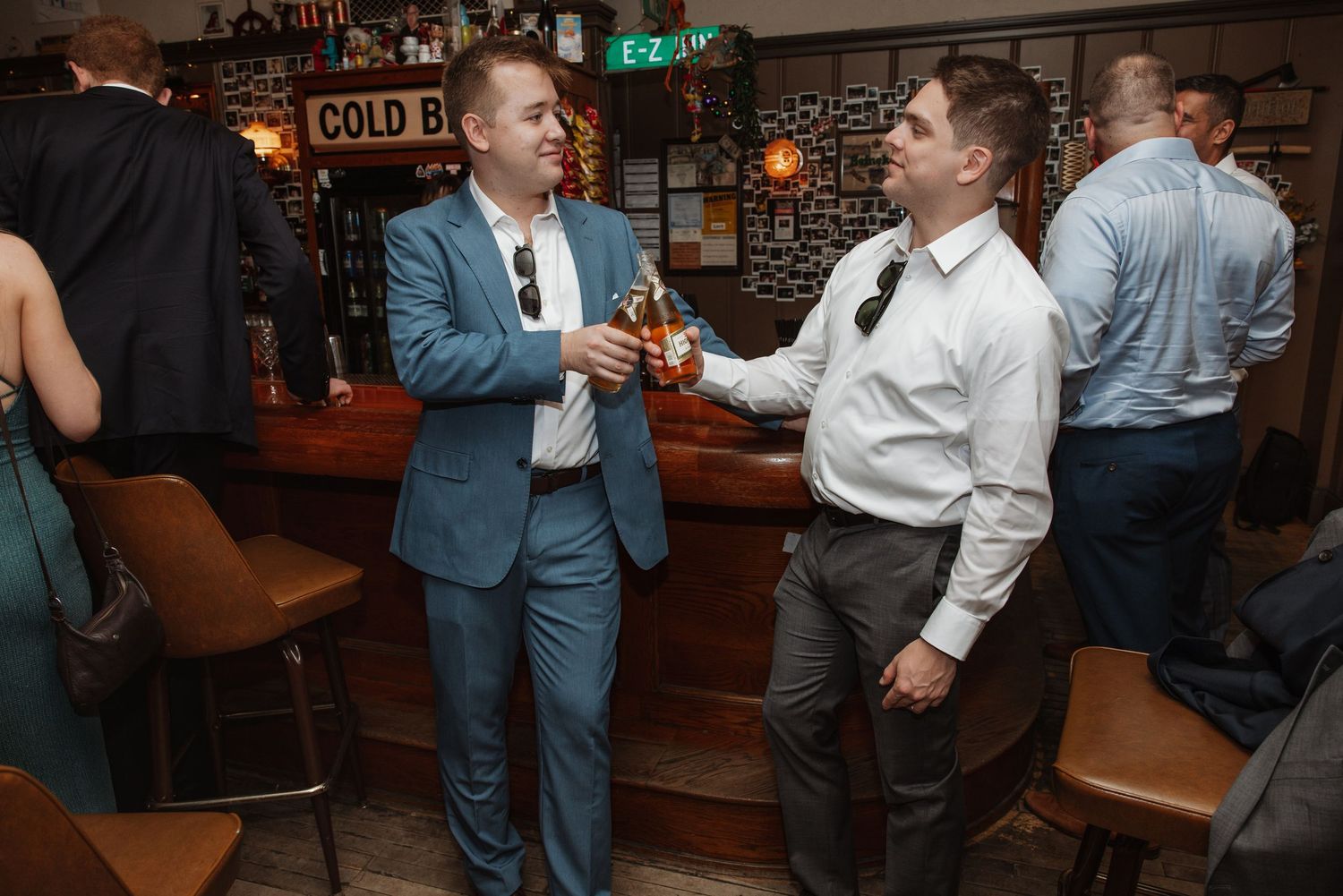 Two men in suits socializing at a bar, clinking bottles in a warmly lit pub setting.