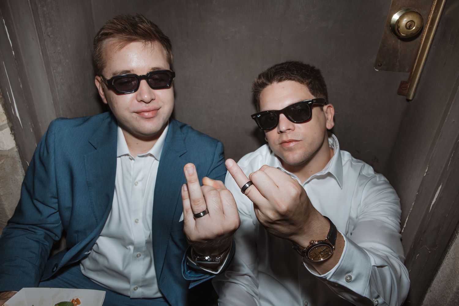 Two men in suits and sunglasses showing off wedding rings while sitting together in a photo booth.