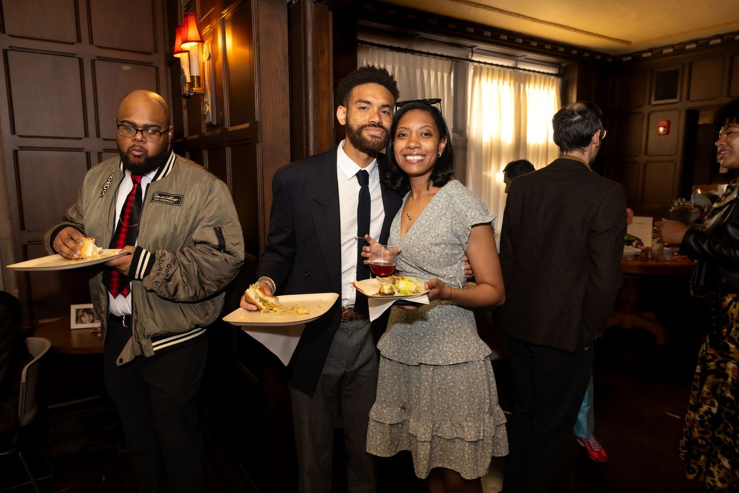 Three guests smile and pose with plates of food at an elegant indoor social gathering event.