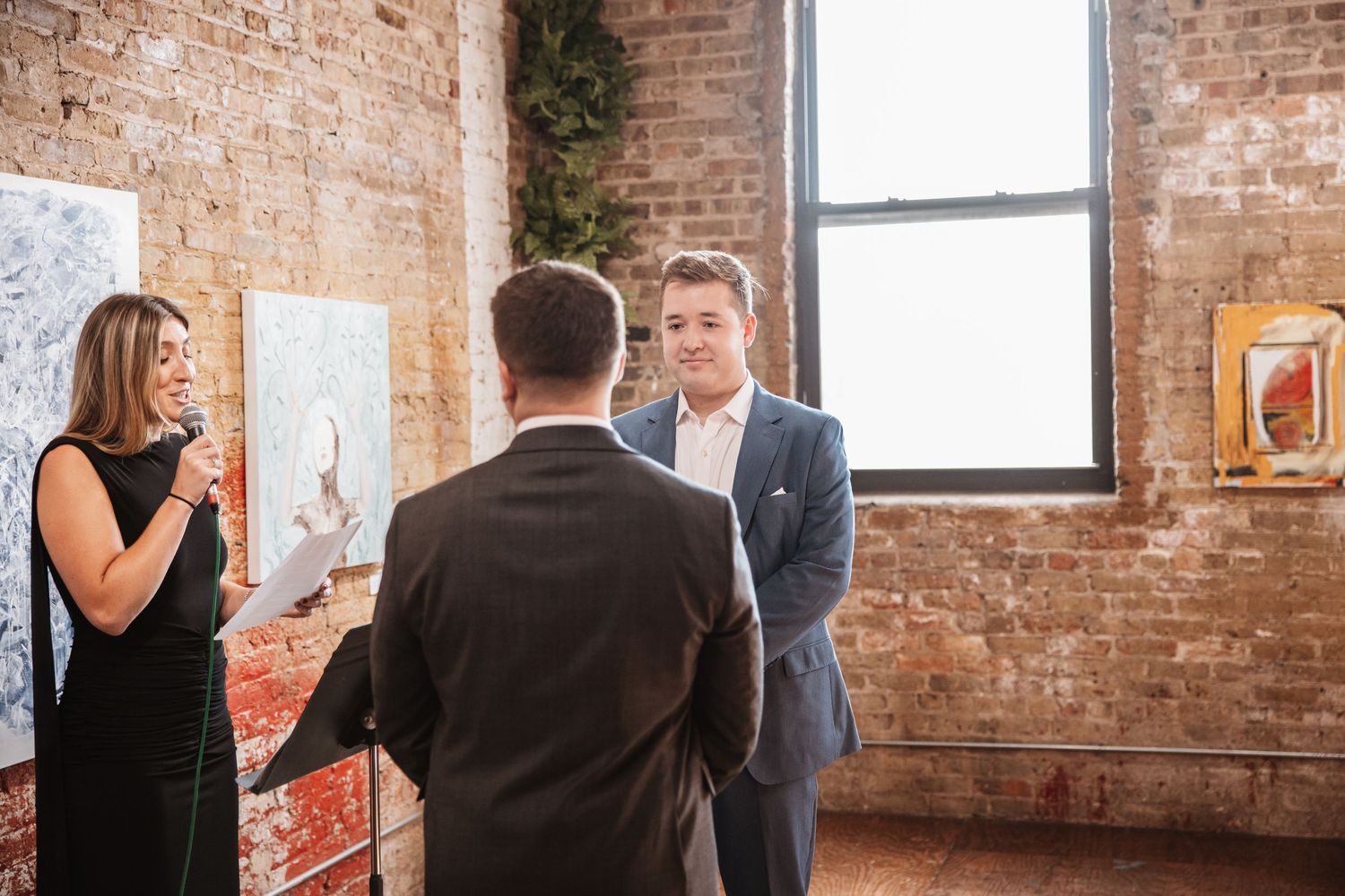 Wedding officiant reading vows as groom in blue suit smiles in art gallery.
