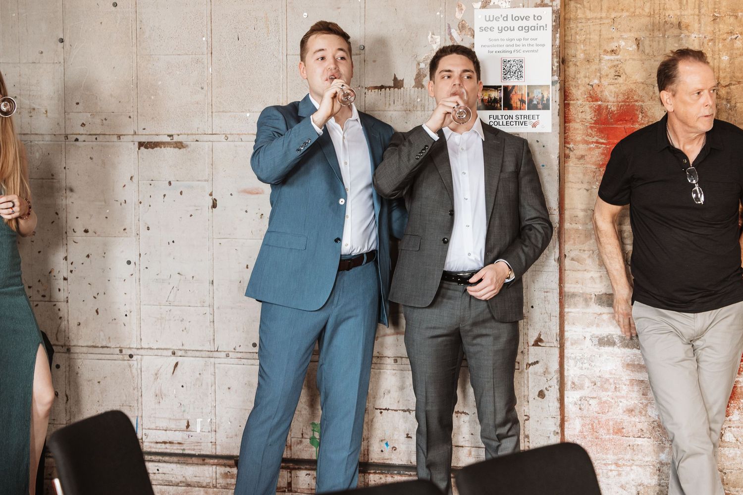 Two men in suits speaking into microphones at an indoor event with brick walls.