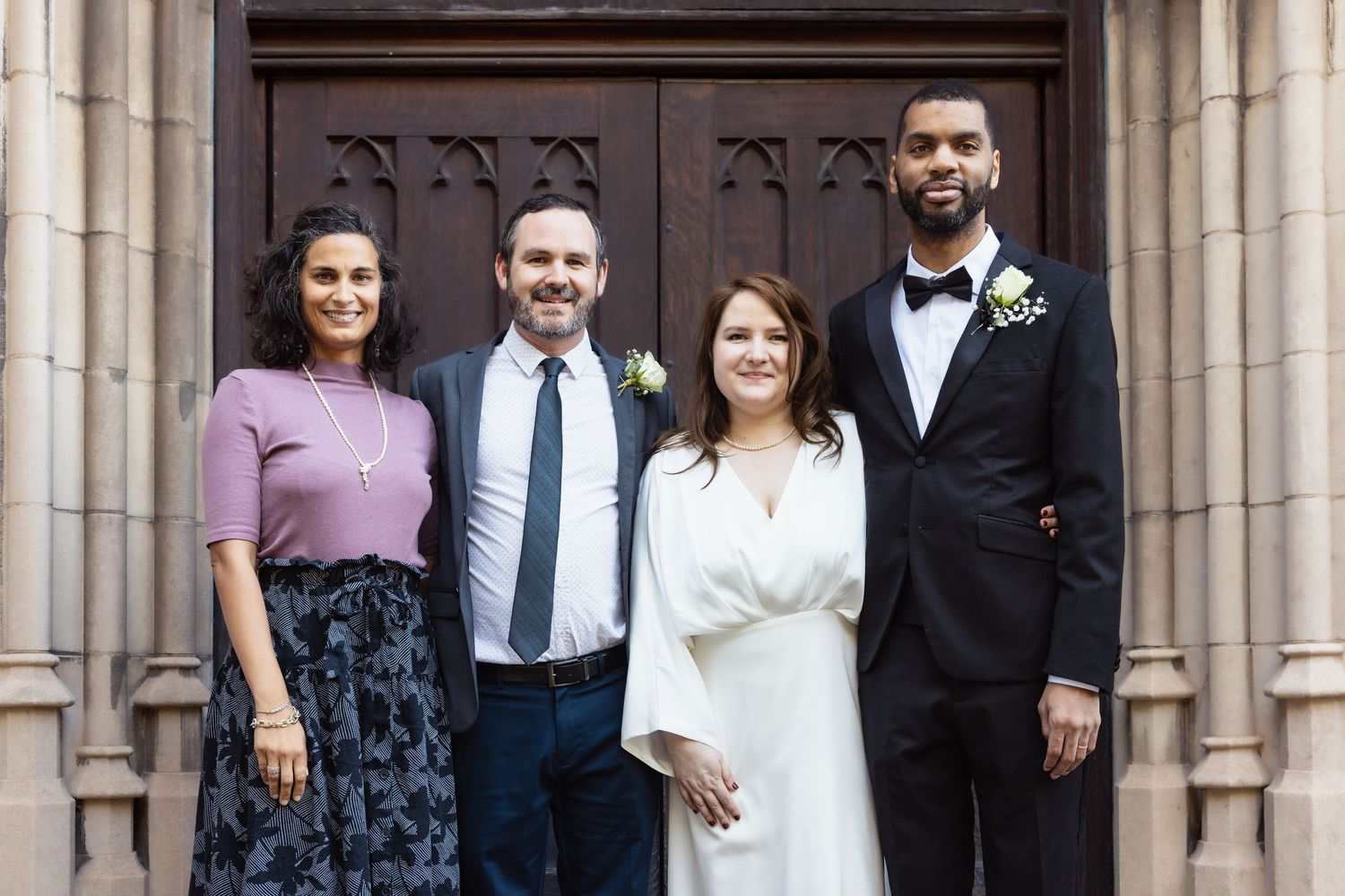 Two couples smile together outside Gothic stone church wooden doors.