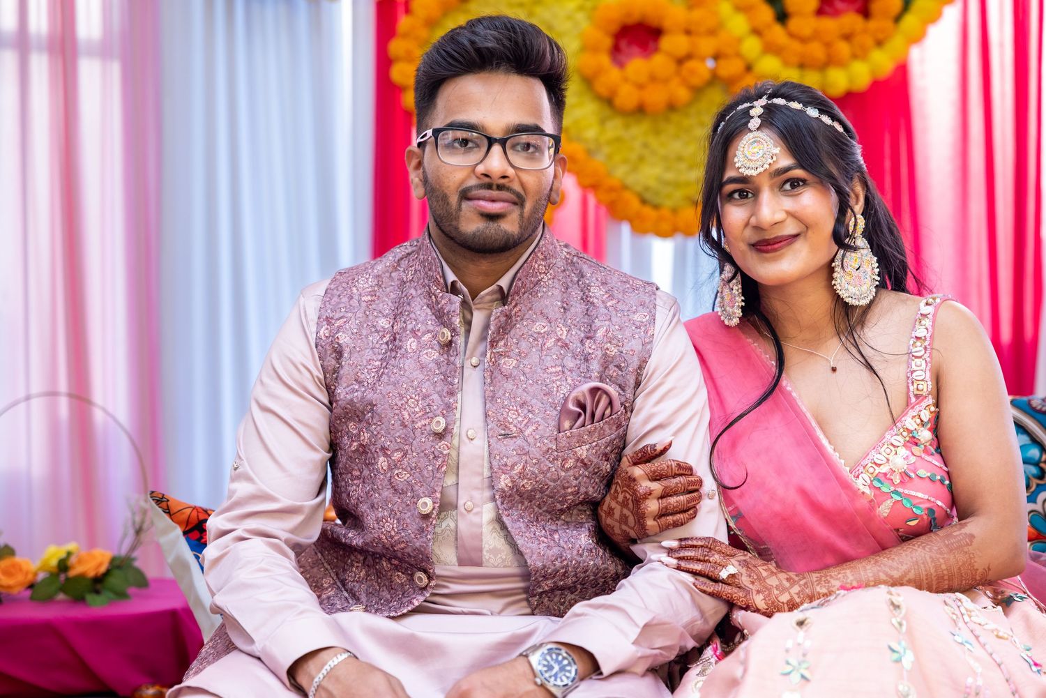 Indian couple in pink traditional attire smiles together at floral mehendi celebration.