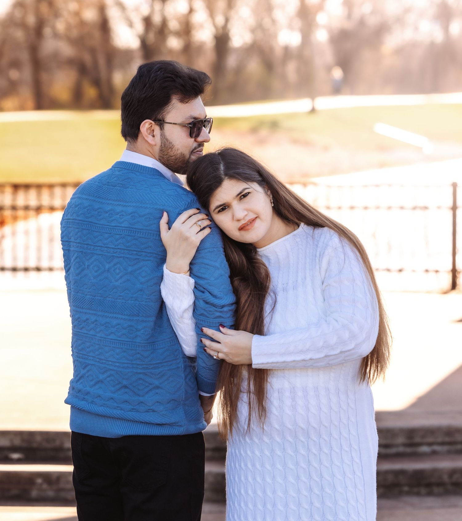 Young couple posing together outdoors with man in blue sweater and woman in white sweater embracing.