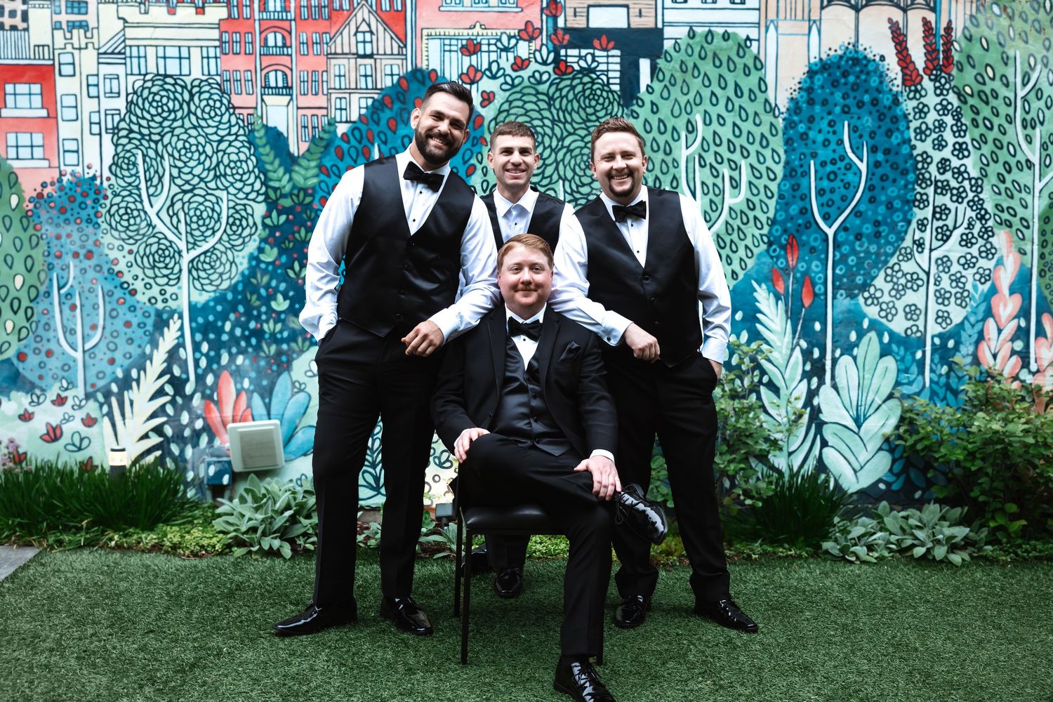 Groom and three groomsmen in black vests posing before colorful garden mural.