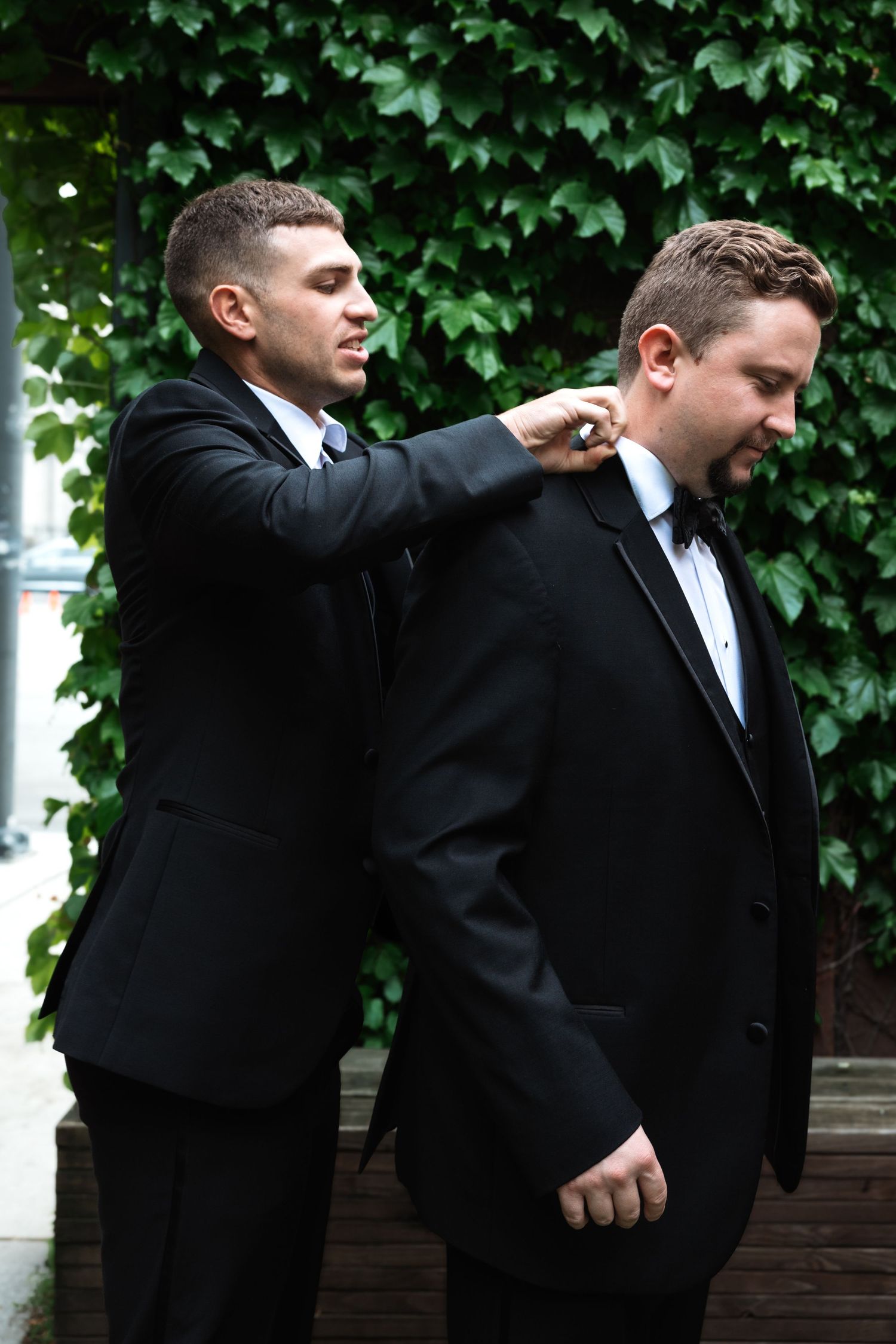 Groomsman in black suit adjusting groom's tuxedo collar outdoors against green ivy backdrop.