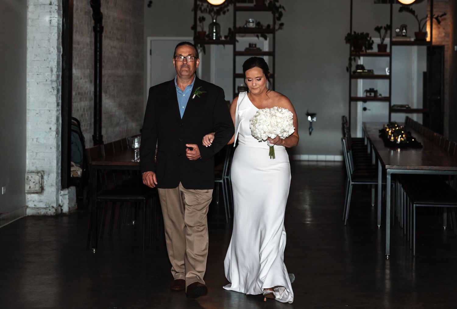 A bride in a fitted white gown walks arm-in-arm with her escort through an industrial wedding venue.