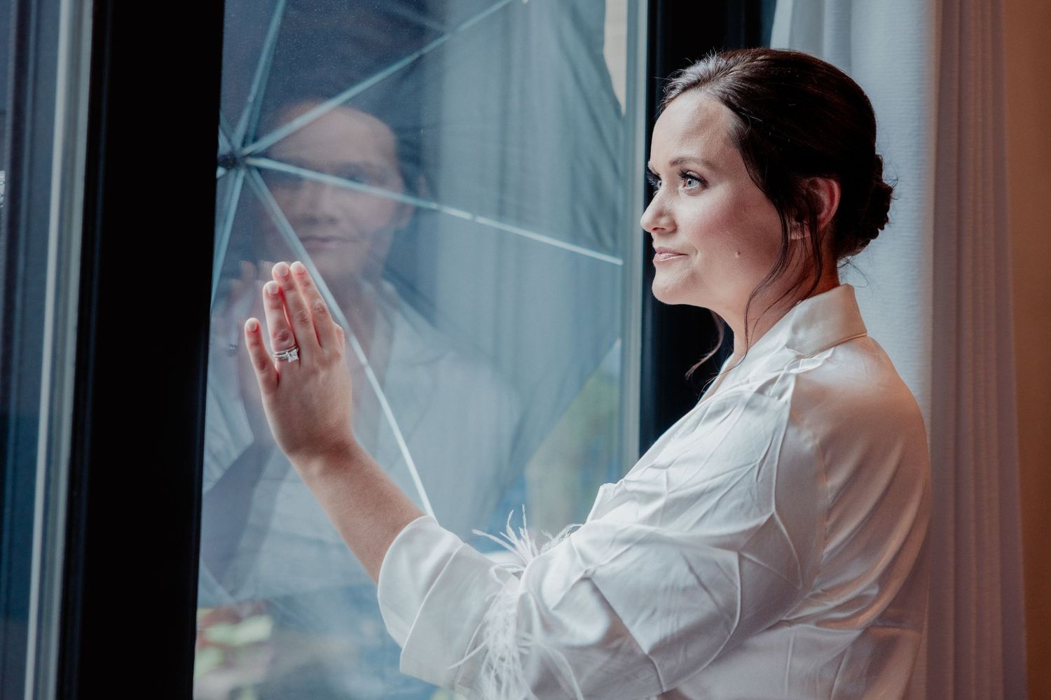 Bride in white robe gazing out window with hand pressed against glass, reflection visible.