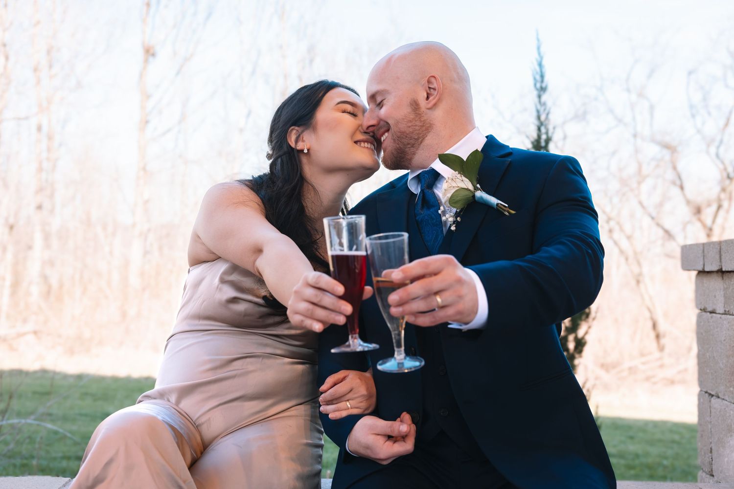 Romantic couple kissing and toasting with red champagne glasses during their outdoor wedding celebration.