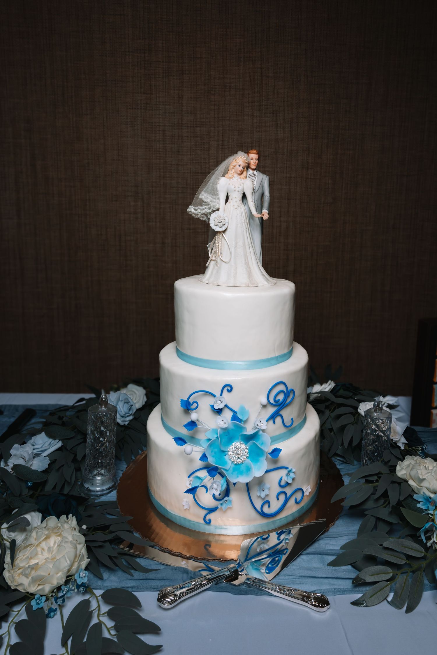 Elegant three-tier white wedding cake with blue floral designs and bride and groom topper.