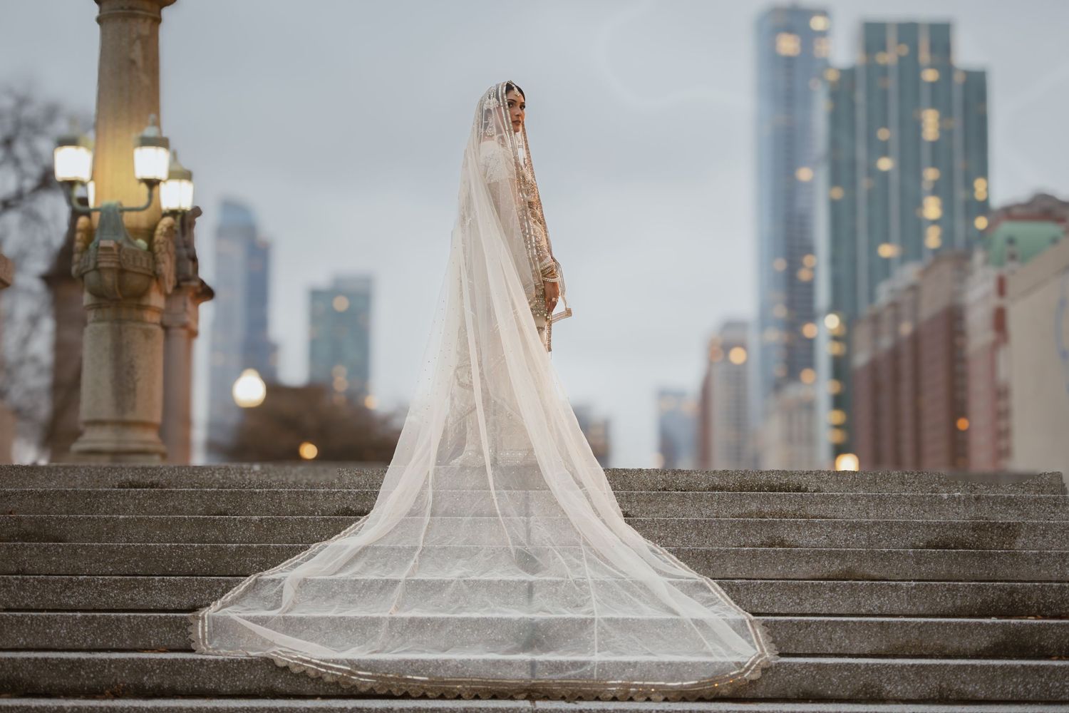 South Asian bride in gold embroidered lehenga with long cathedral veil on steps with city skyline.