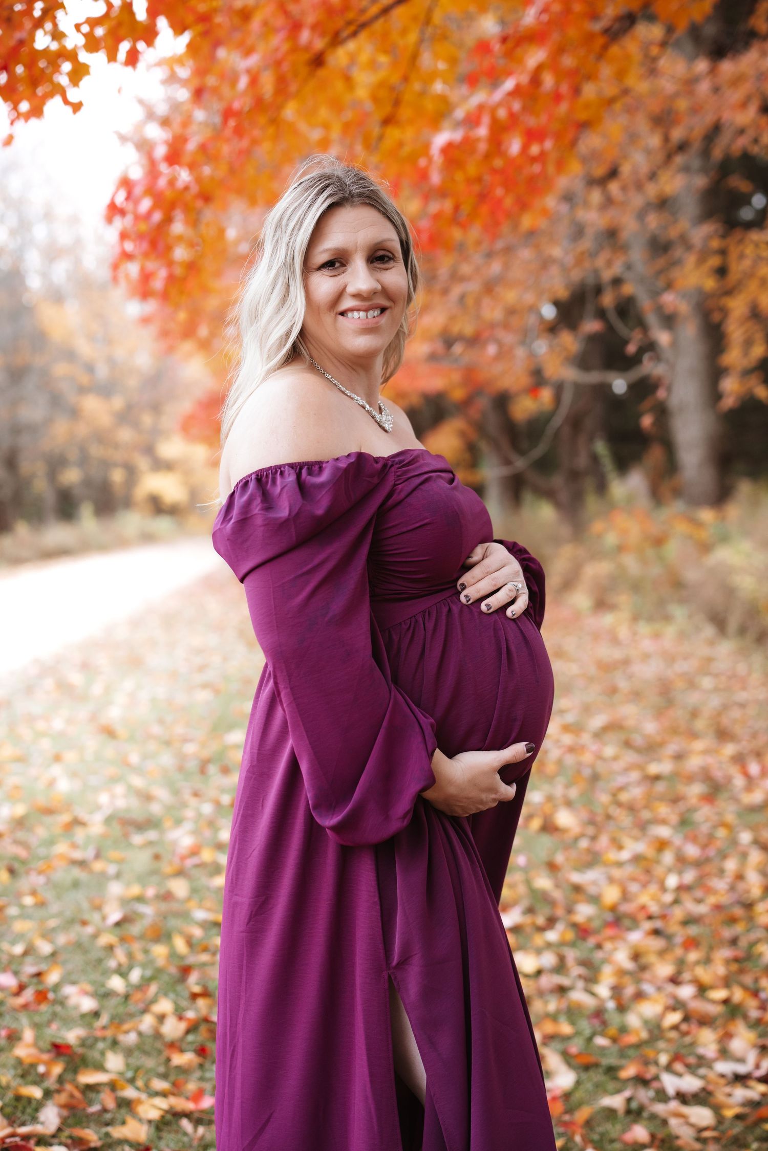 Happy pregnant woman in purple off-shoulder gown embracing baby bump among stunning fall foliage.