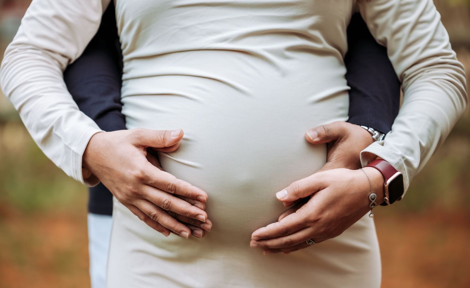 Close-up of a pregnant woman in white dress with partner's hands cradling her baby bump outdoors.