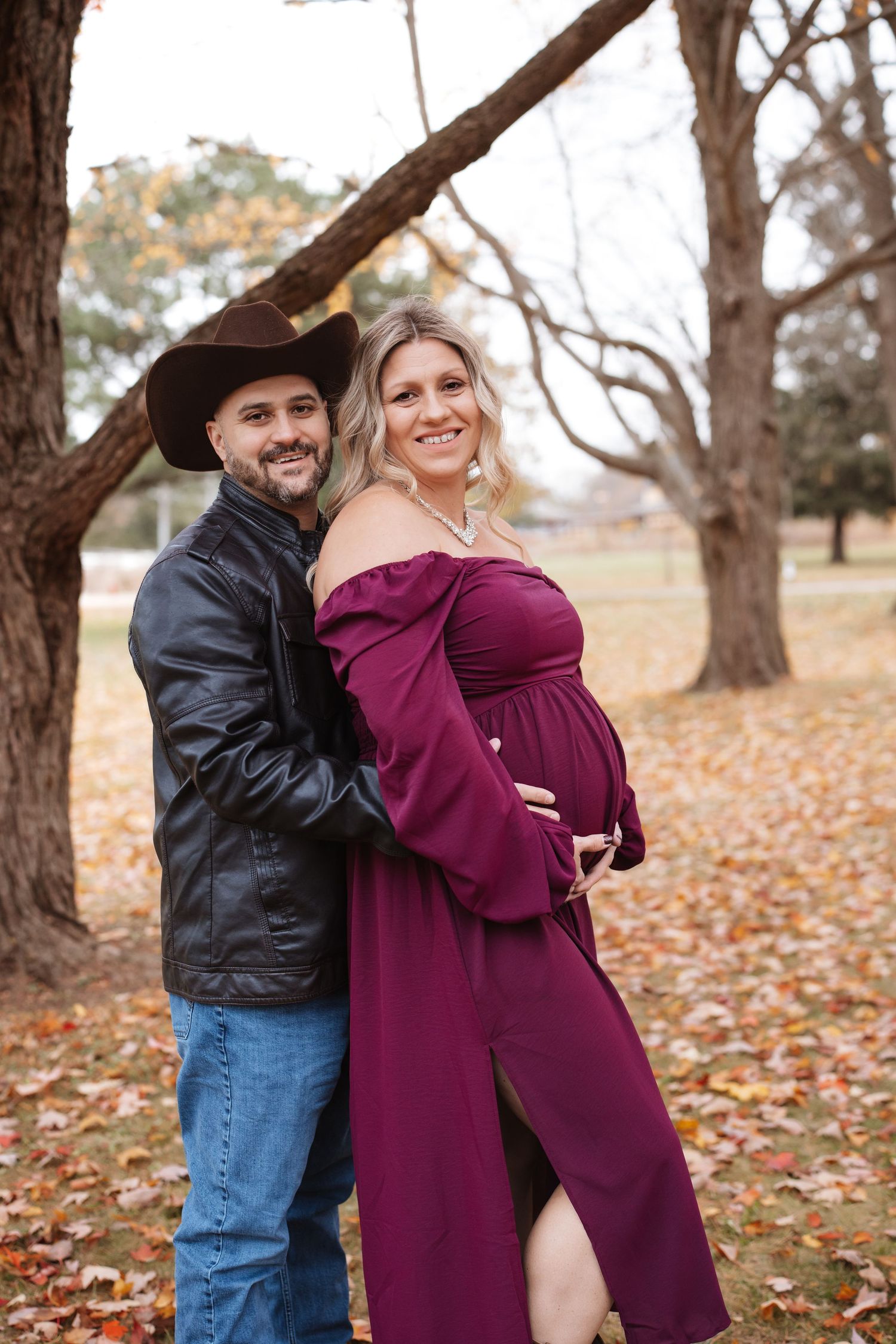 Smiling couple in fall foliage park, man in cowboy hat hugging pregnant woman in off-shoulder burgundy maternity gown.
