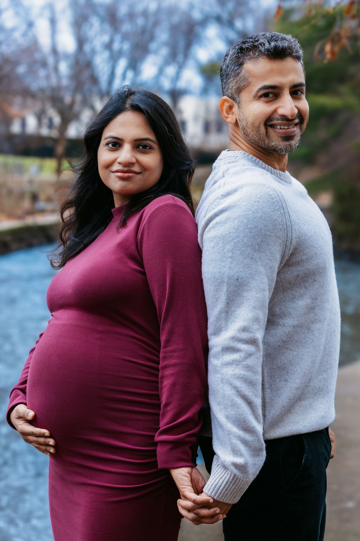 Pregnant couple standing back-to-back holding hands, smiling at camera near a serene park waterway.