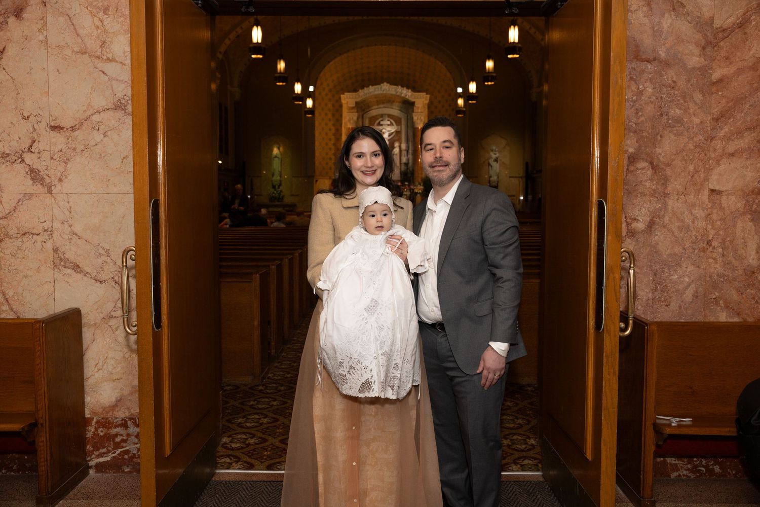 Family posing with a baby in a white christening gown at the entrance of a Catholic church.