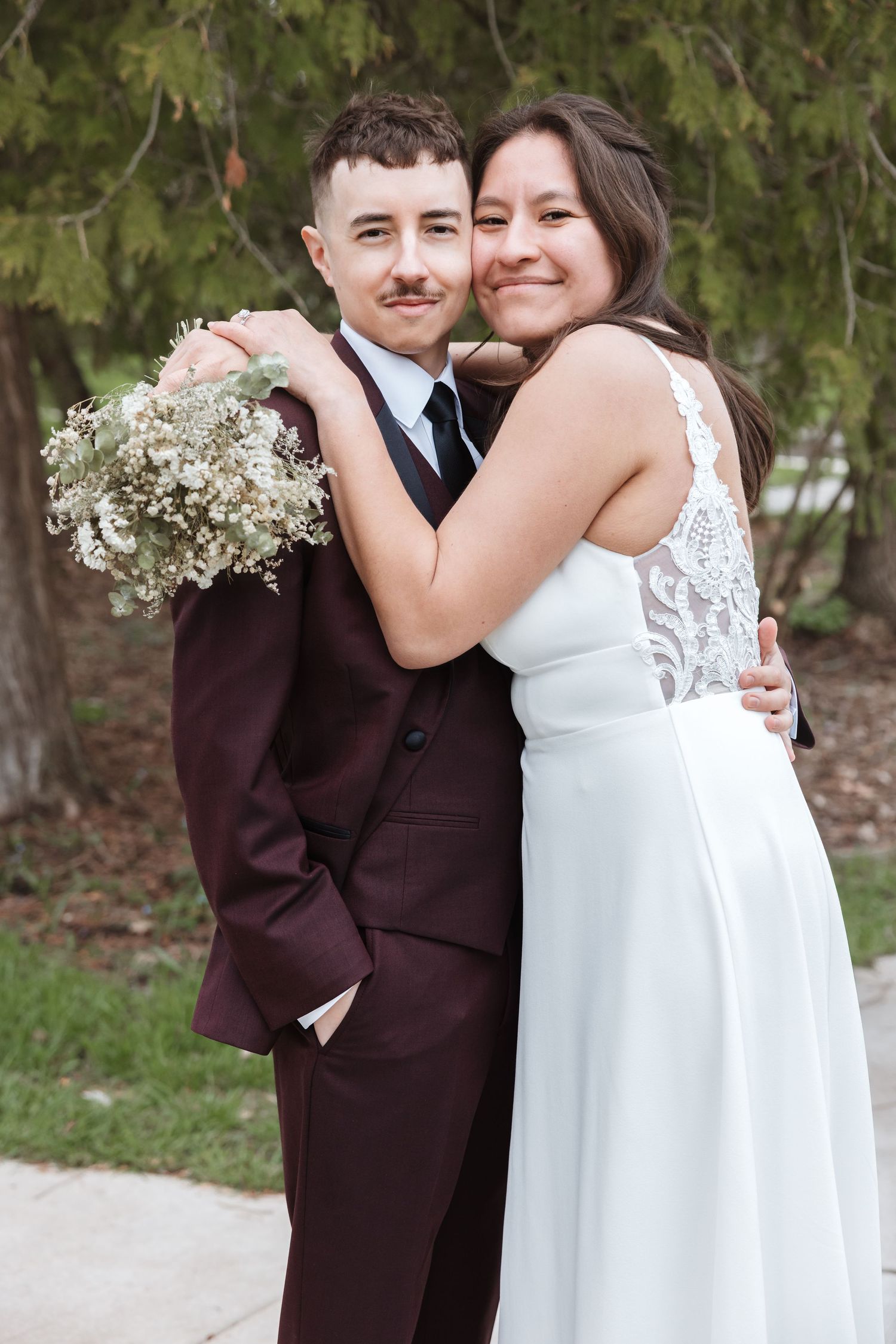 Bride in white lace dress hugs groom in burgundy suit holding bouquet outdoors at wedding ceremony.