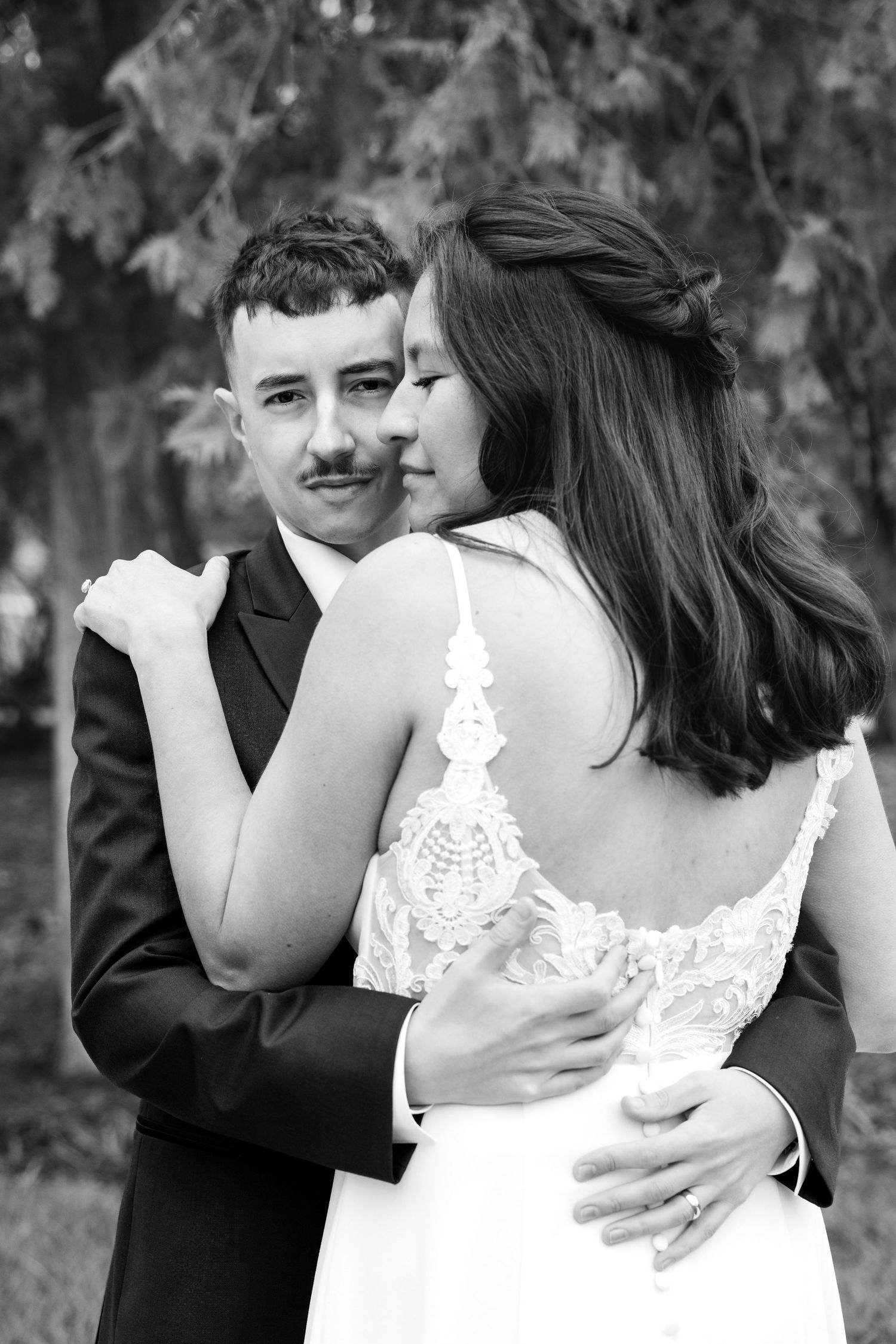 Black and white wedding photo of bride and groom embracing outdoors, bride in lace dress, groom in suit with mustache.