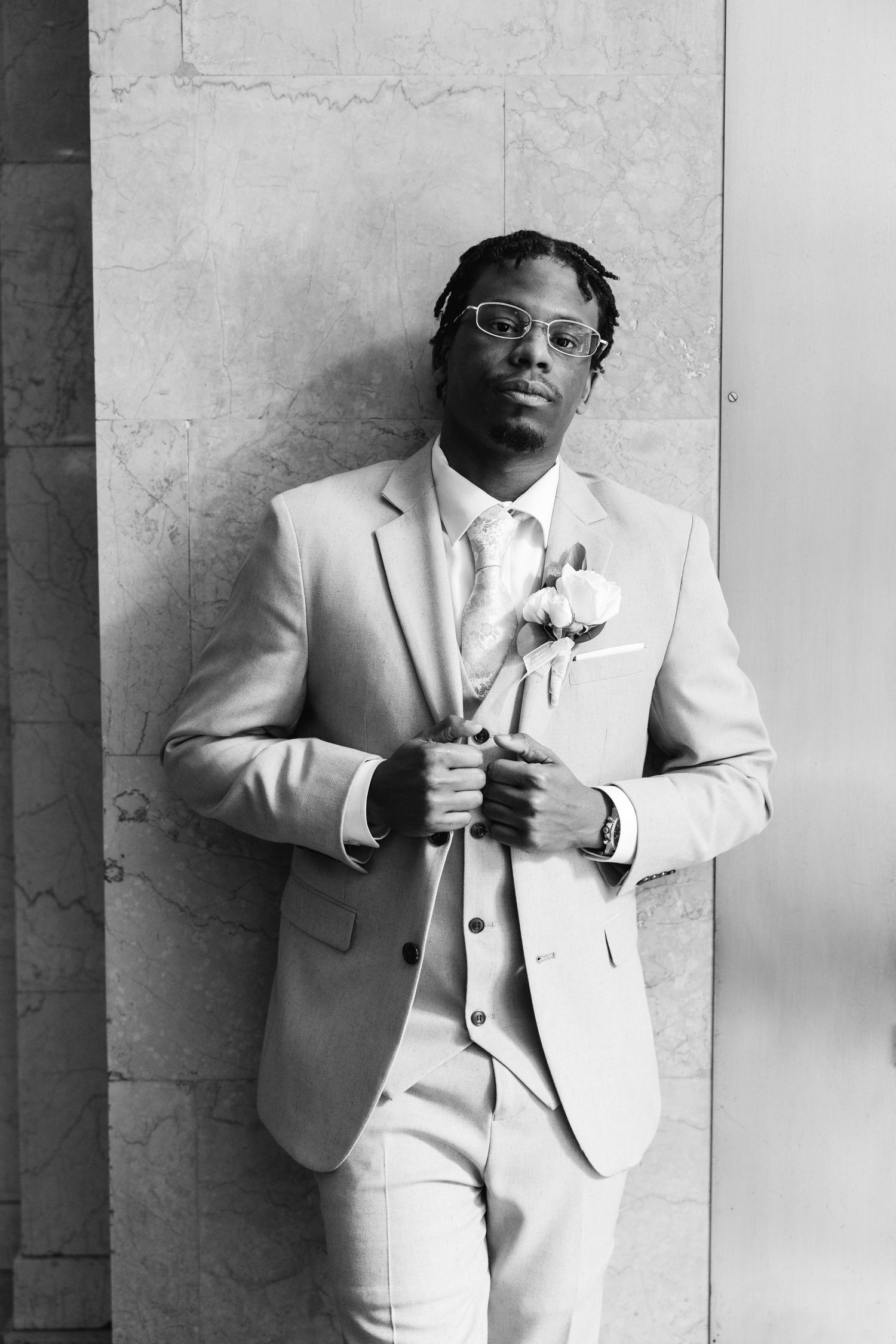 Groom in light gray three-piece suit with boutonniere poses against marble wall in black and white portrait photo.