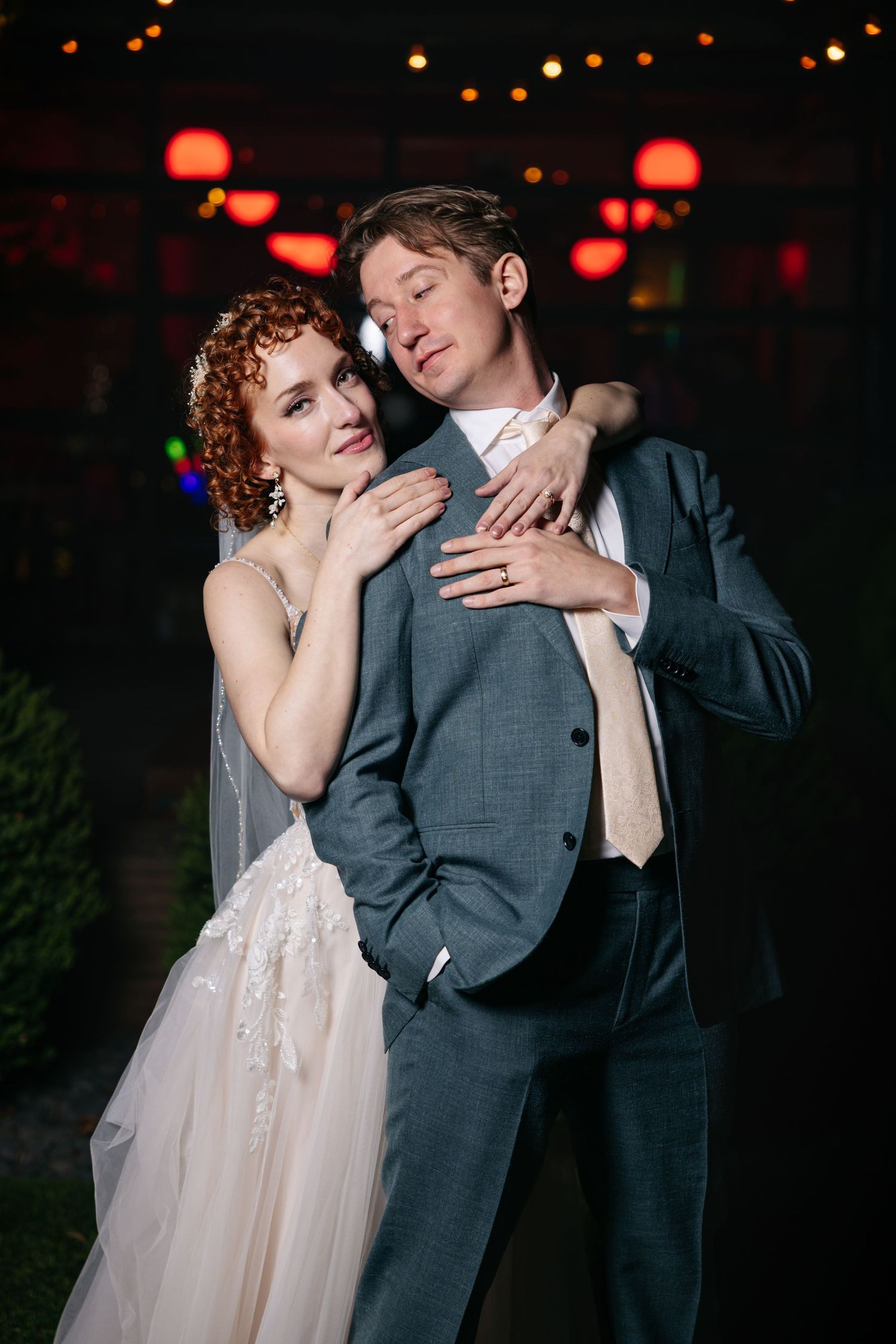 Red-haired bride in white gown embraces groom in gray suit against dark background with red lanterns.